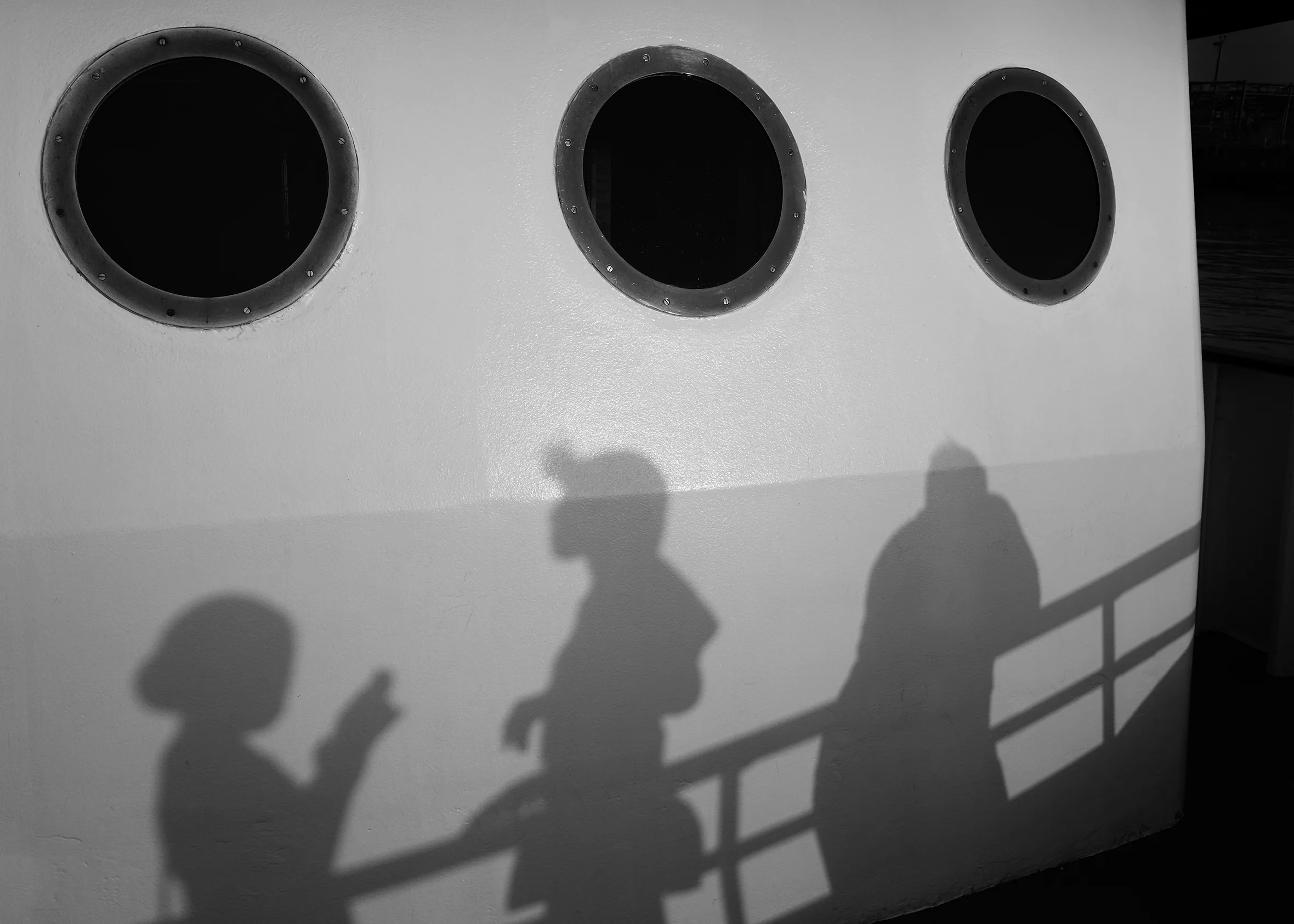 A close-up of three portholes on a ship. Reflections of three people against an open railing are on the side of the ship underneath the portholes.
