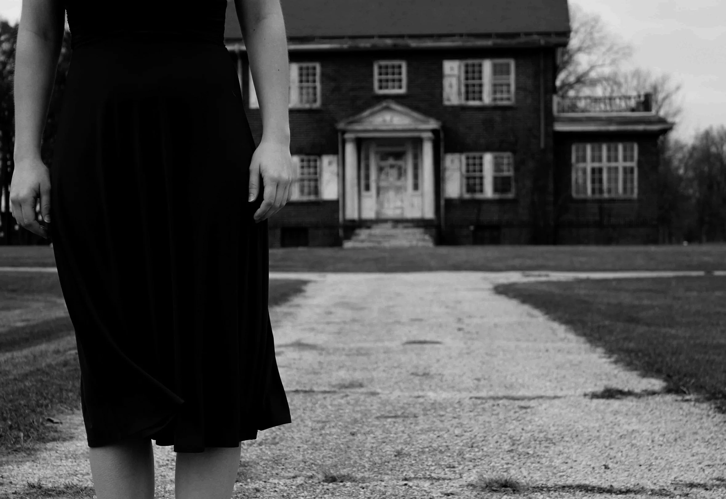 A woman in a dress and facing the camera is seen from the midriff to below the knee with her arms straight down. She is standing on a sidewalk at a distance from the front of an old two-story house.