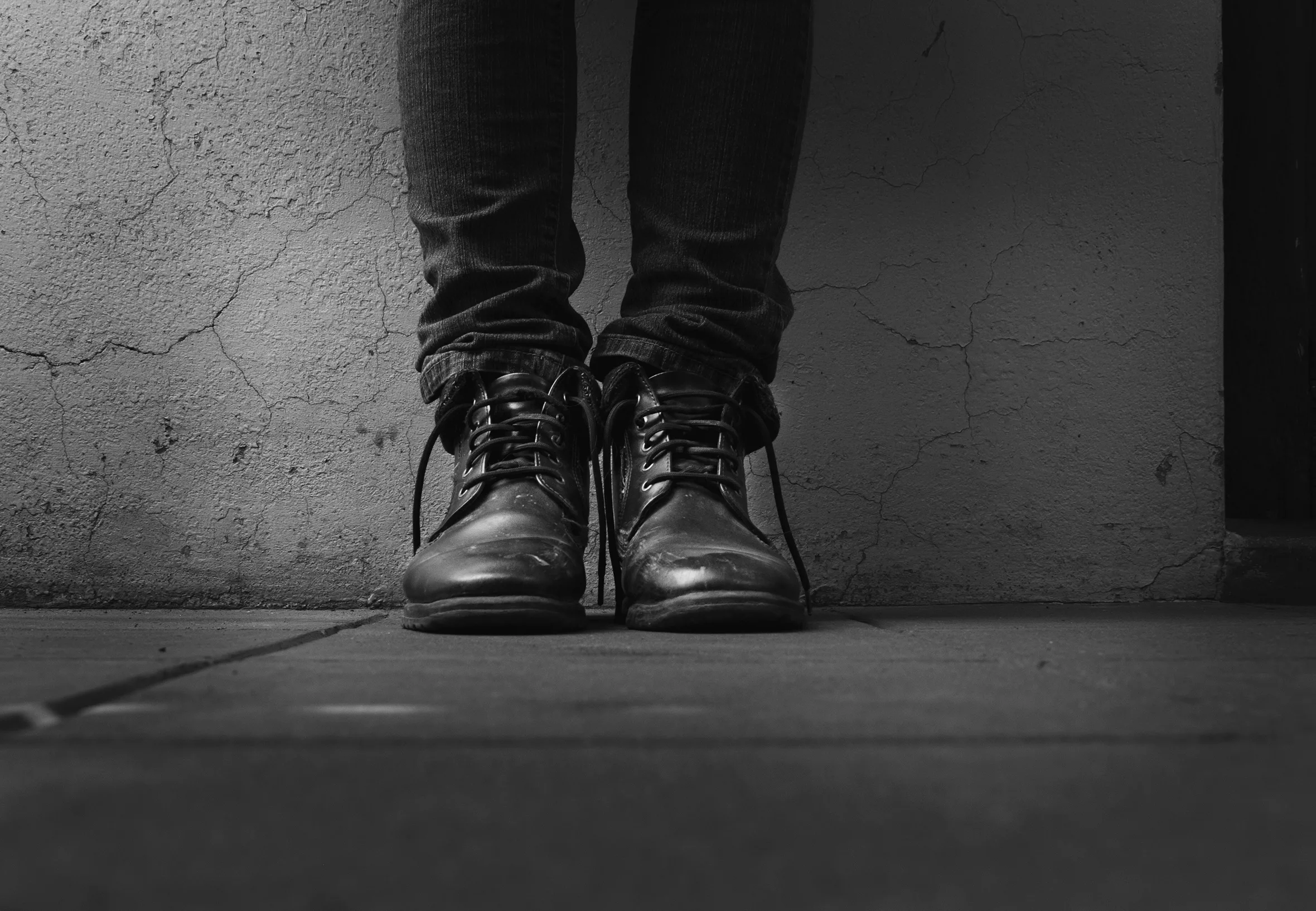 A person’s legs shown below the knees standing against a painted, concrete wall with cracks in it. The person is wearing fitted jeans scrunched above ankle-height, laced boots that are untied with the laces hanging down.