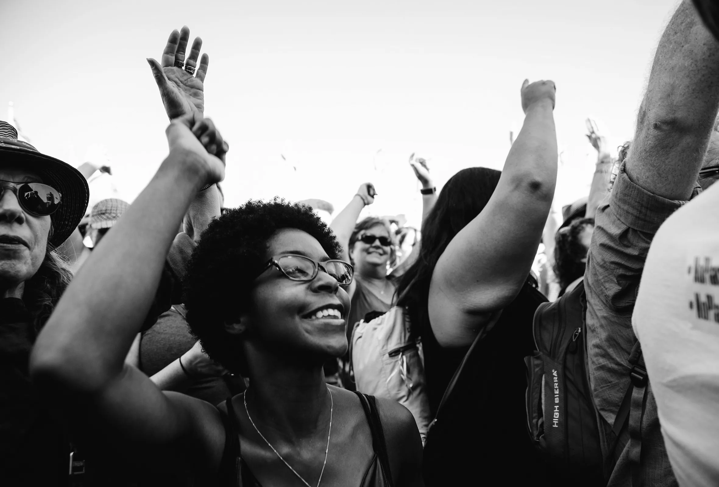 A Black woman, smiling, throwing her hand up in the air among a diverse crowd of protesters.