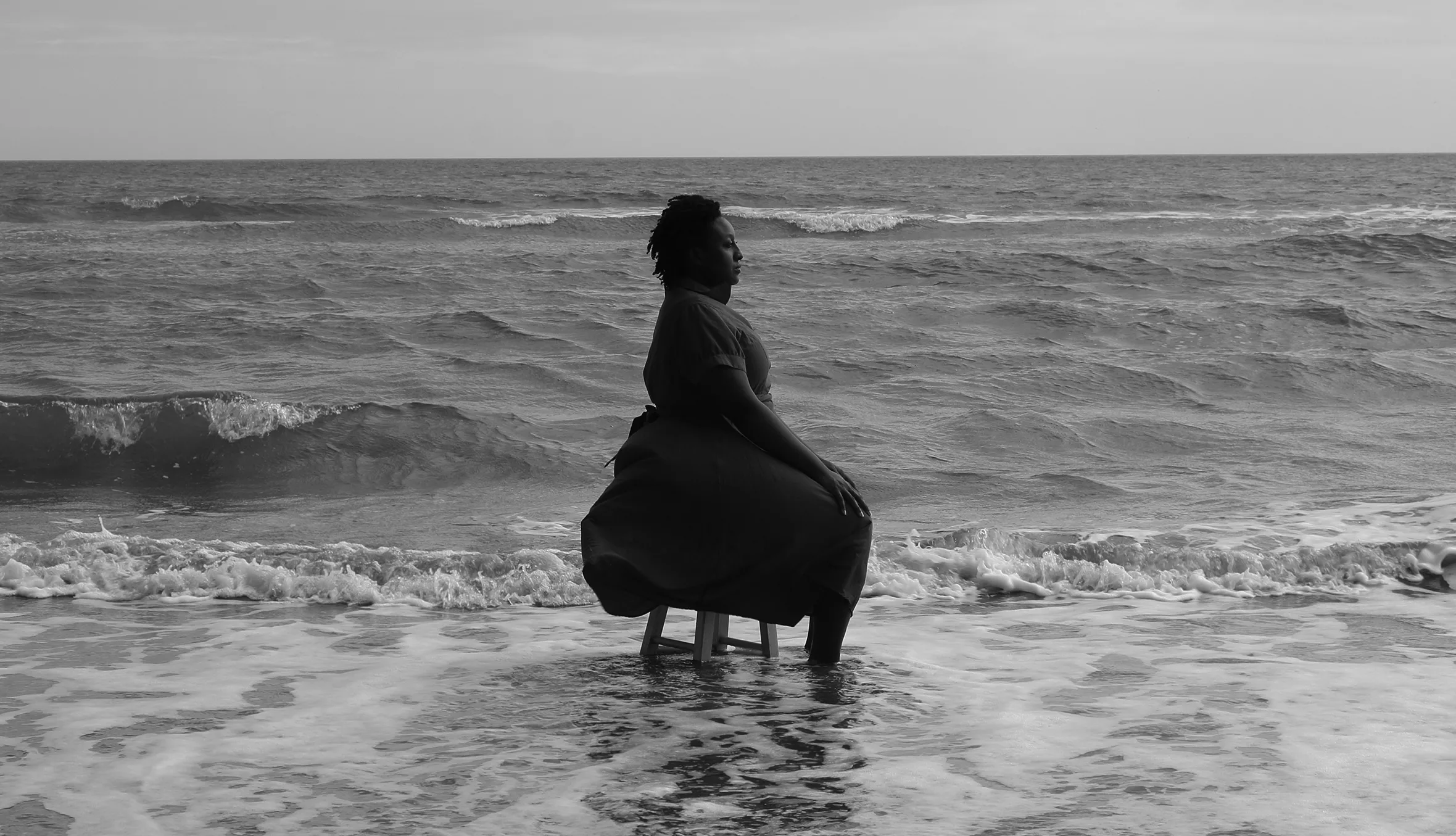 A woman in a dress sits in profile on a short stool in the surf on the beach on the Isle of Palms, South Carolina. The waves are mild and the water is midcalf level.