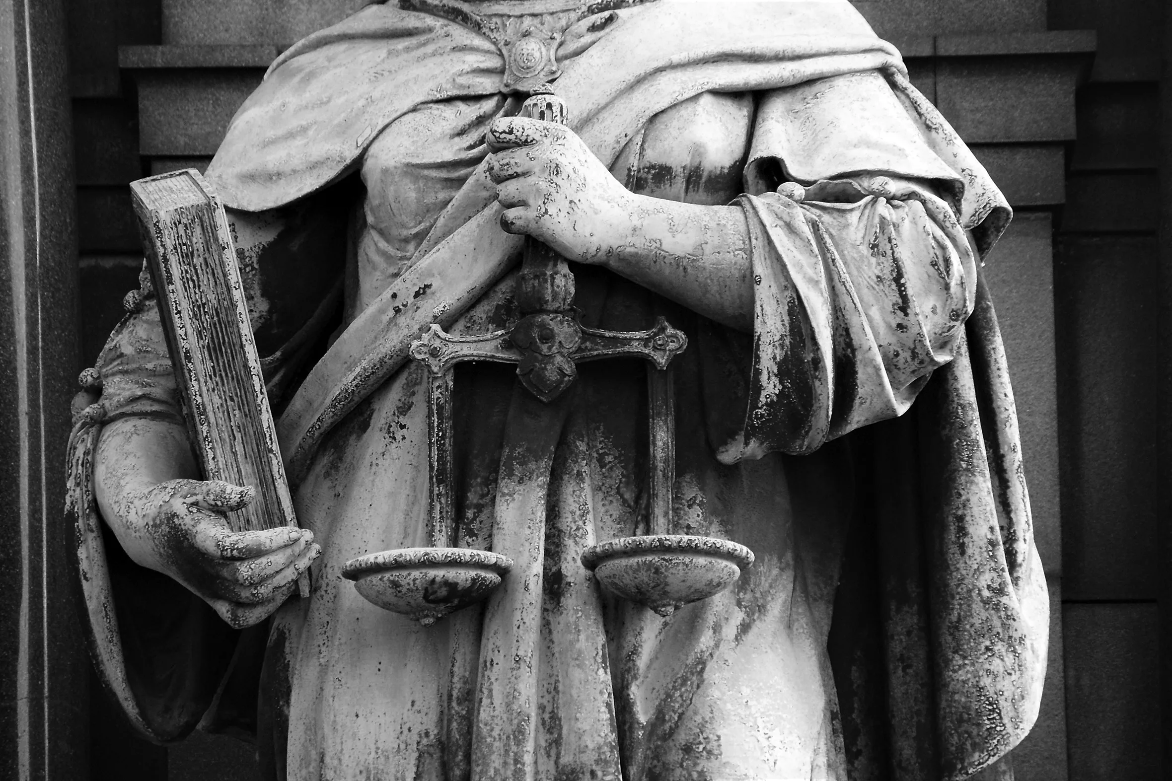 A large statue of a woman holding a book and a balance scale dressed in a gown with a cape is shown from shoulder to midthigh.
