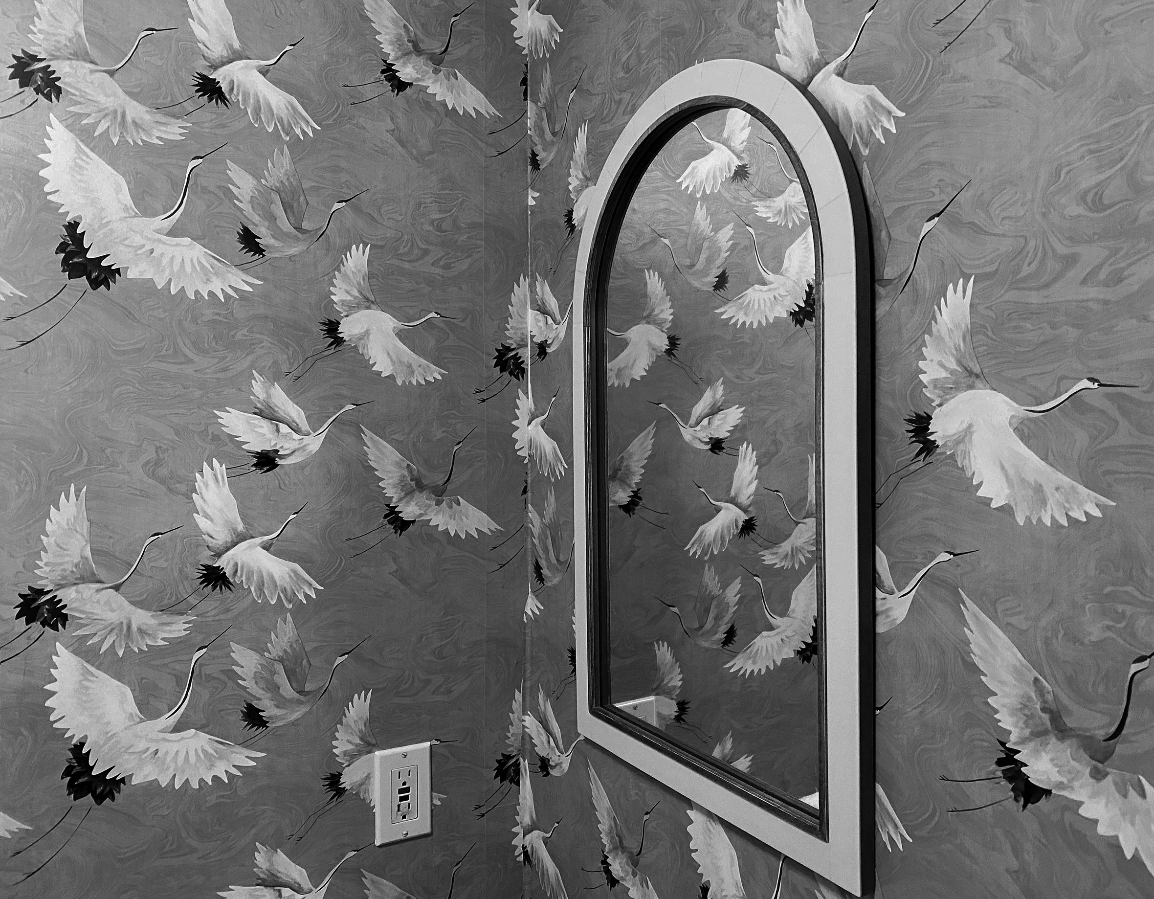 An arched mirror next to a corner with wallpaper full of flying cranes.