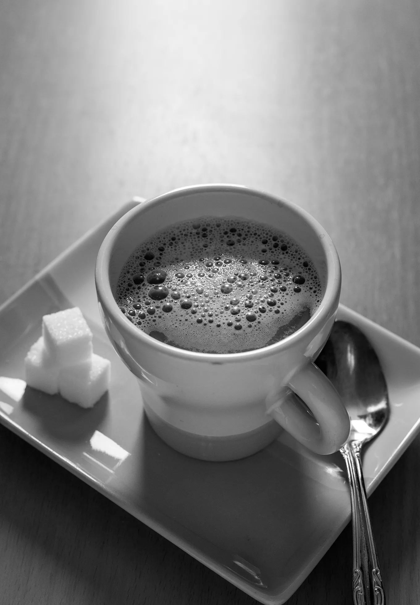 Close-up of a cup of coffee on a rectangular serving plate. Three sugar cubes are stacked to the left of the cup and a teaspoon is placed to the right.