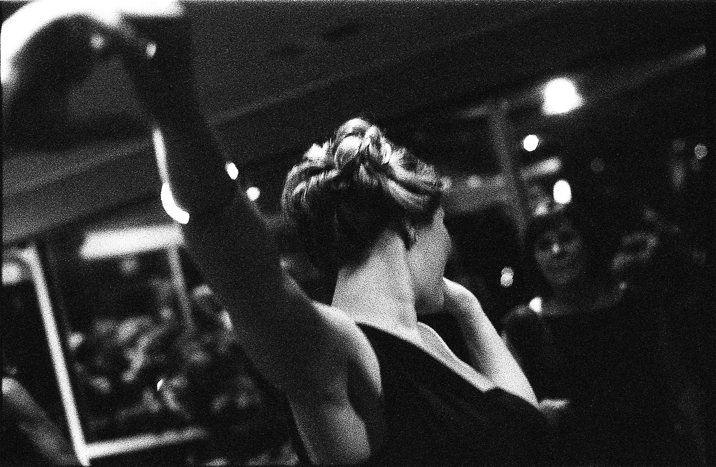 A woman with an elegant, updo hairstyle is facing away from the camera. She appears to be dancing in a darkened room. The shadows of people can just be seen around the edges.