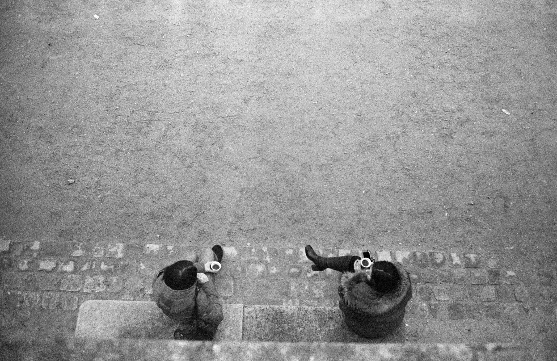 Two women in heavy coats sit angled toward one another in conversation on a concrete bench while drinking coffee from white styrofoam cups.
