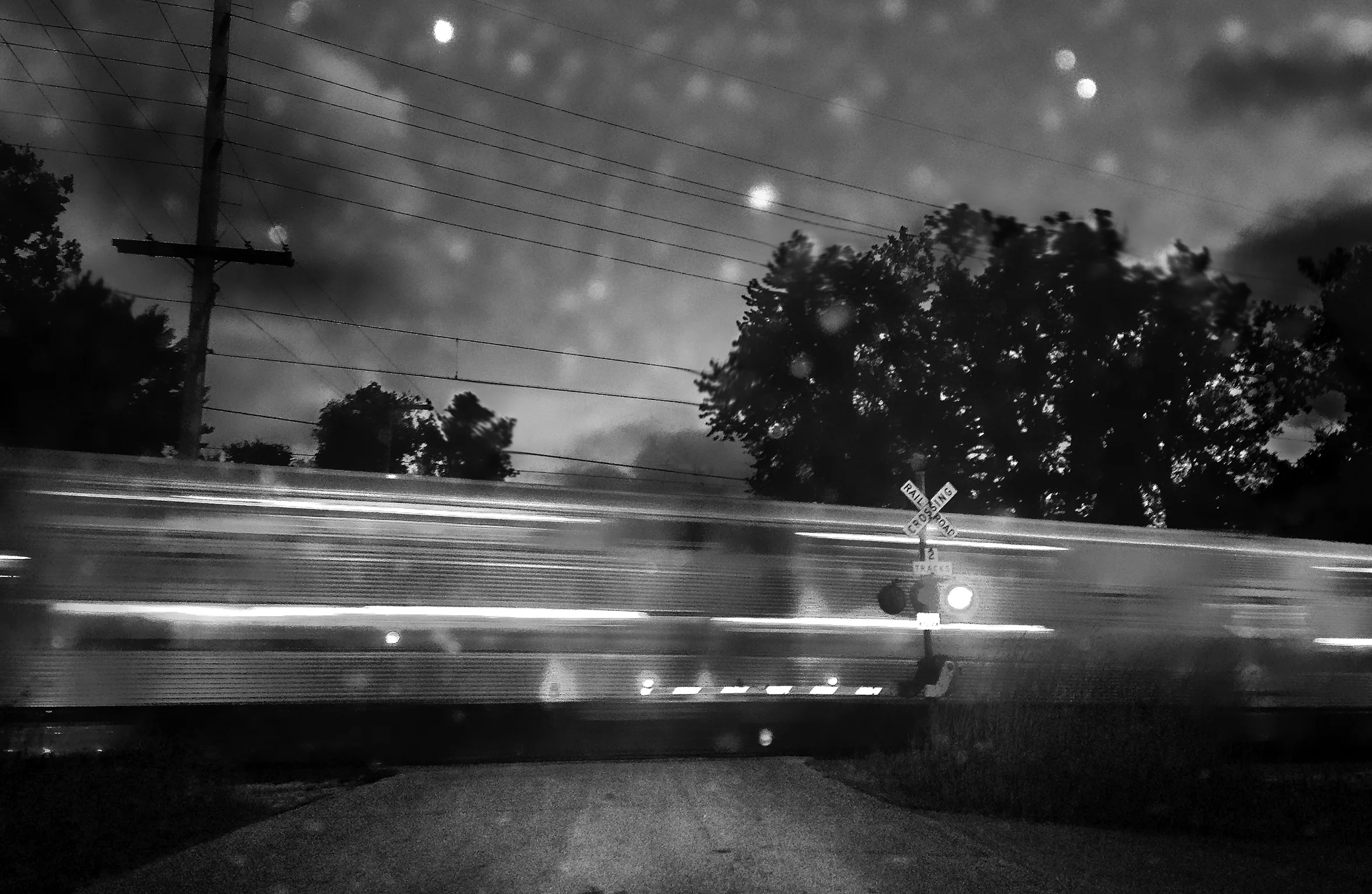 A train whizzes by a railroad crossing. Light is bouncing off the moving train and coming from the railroad crossing signal and seen pixelated throughout the sky.