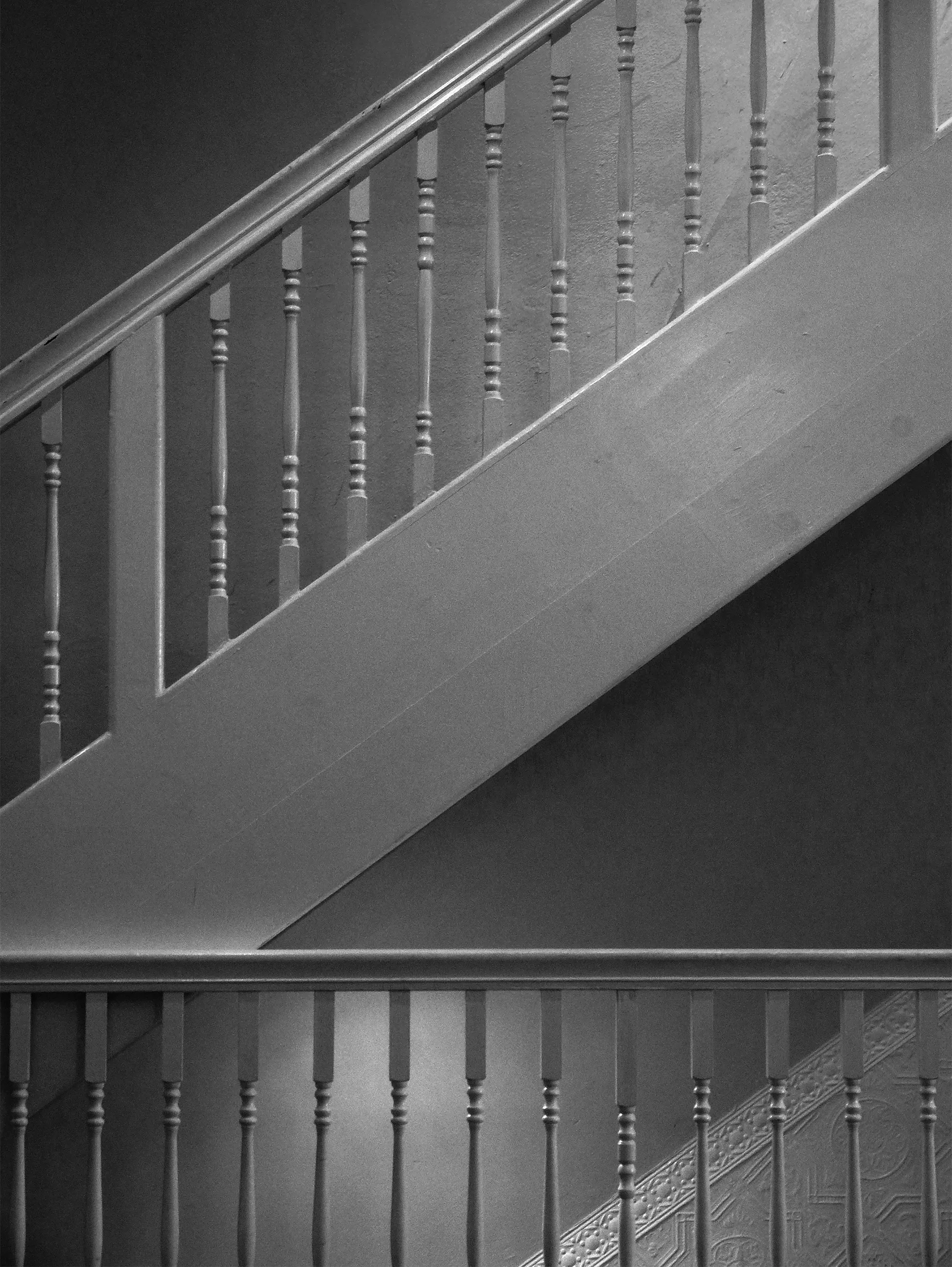 The upper part of wooden balusters and handrail of a second floor walkway in front of a matching wooden staircase leading to the third floor.