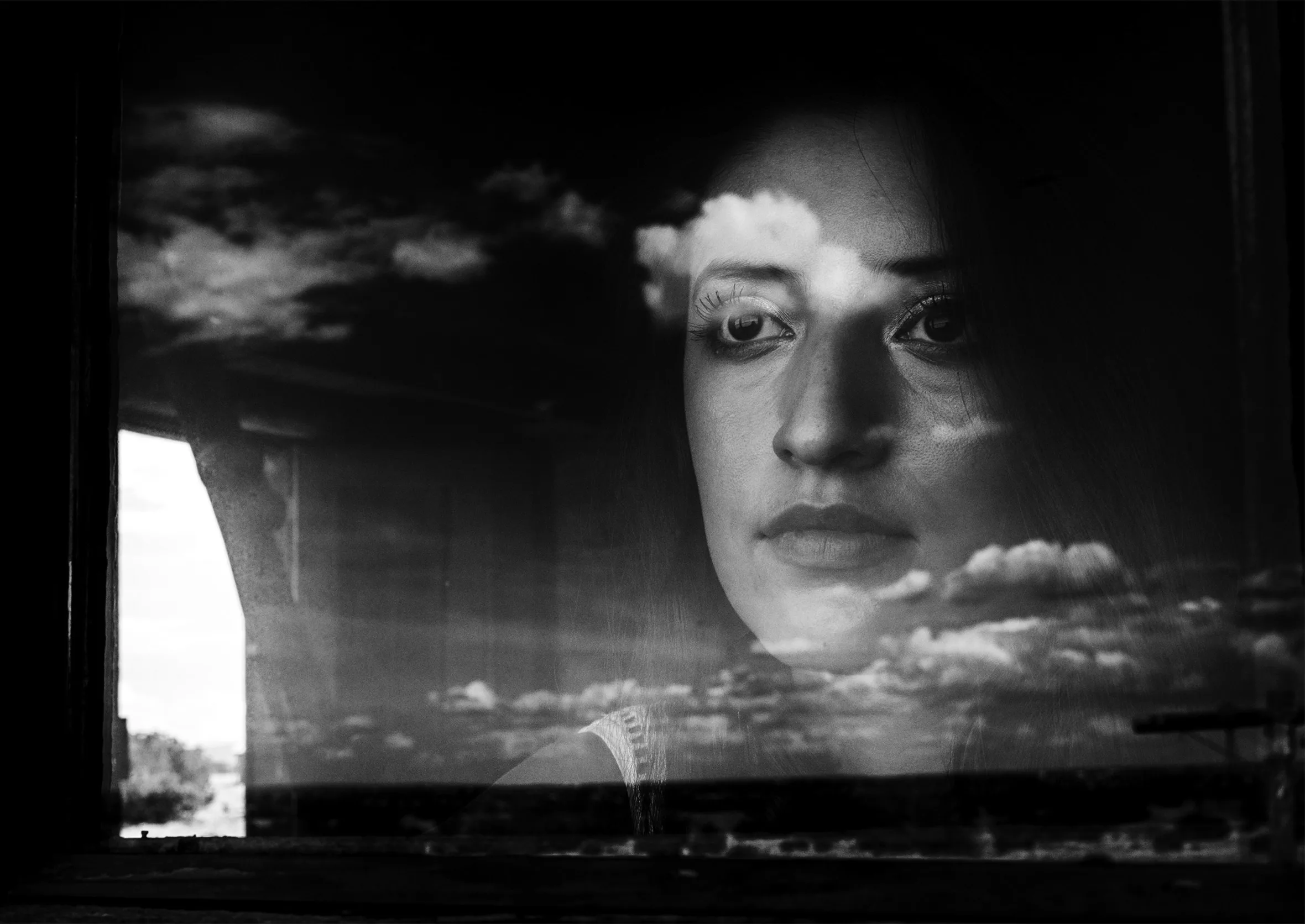 A young woman with dark hair looks off into the distance from behind a dark-framed window. The room’s interior is visible through the reflection of the outside vista and white clouds on the windowpane.