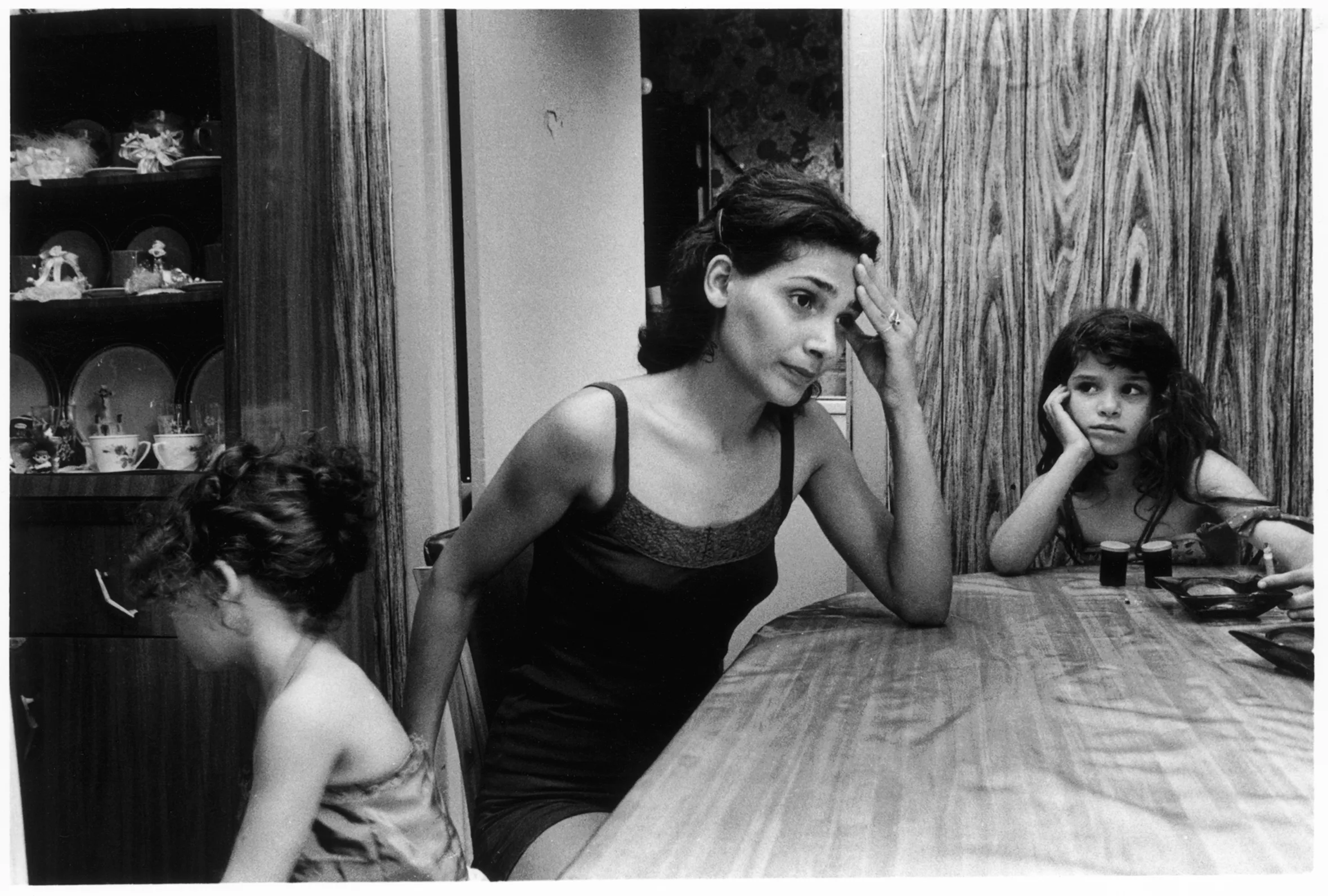 A frustrated-looking woman sits at a dining-room table with two young girls. The woman has her left elbow on the table with her hand on her forehead. One girl faces away, while the other rests her head in her hand and looks at the woman.