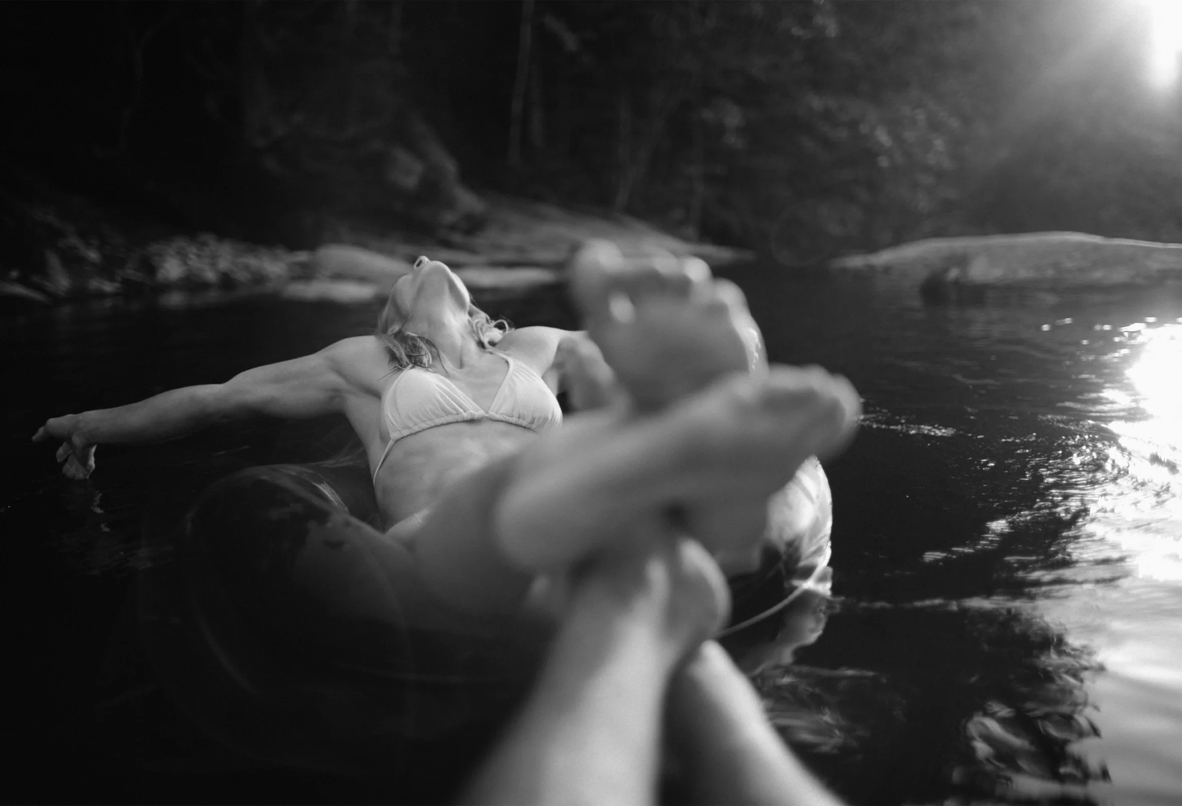 A woman in a bikini with outstretched arms and head tilted back lounges on an inner tube. Her feet in the foreground, soles facing the camera, are intertwined with another’s, who is mostly off camera, as they float down a river on inner tubes.