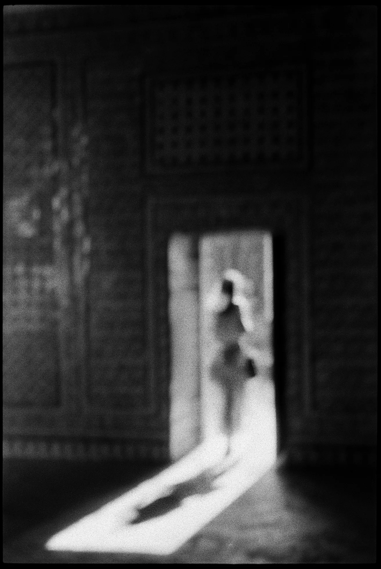 Blurry photo of a woman walking through a large, rectangular opening in a structure into the bright sunlight with her long shadow trailing behind.