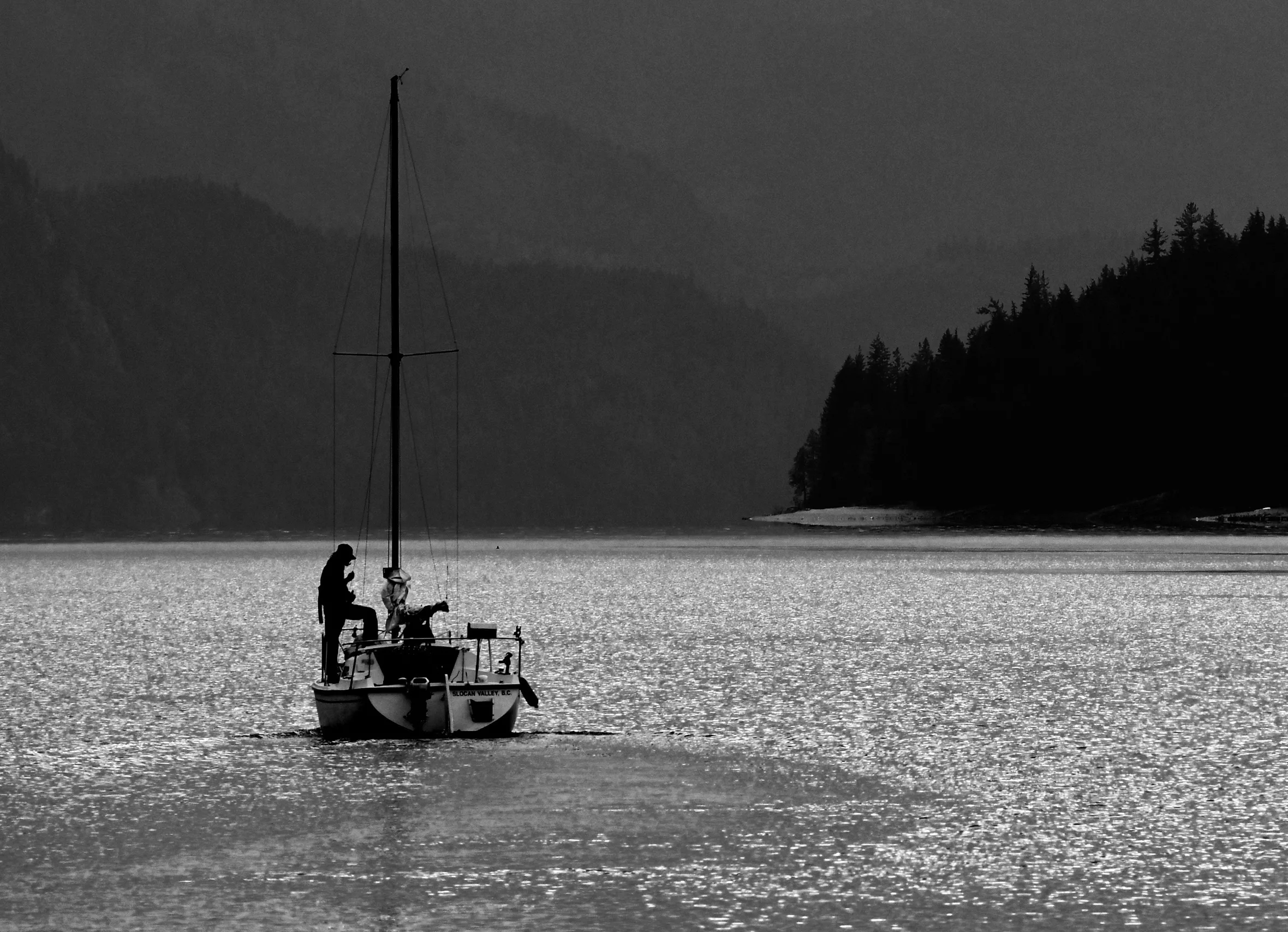 Sailboat at sunset with a person in silhouette on deck and the shoreline of Slocan Lake in British Columbia, Canada, in the background.