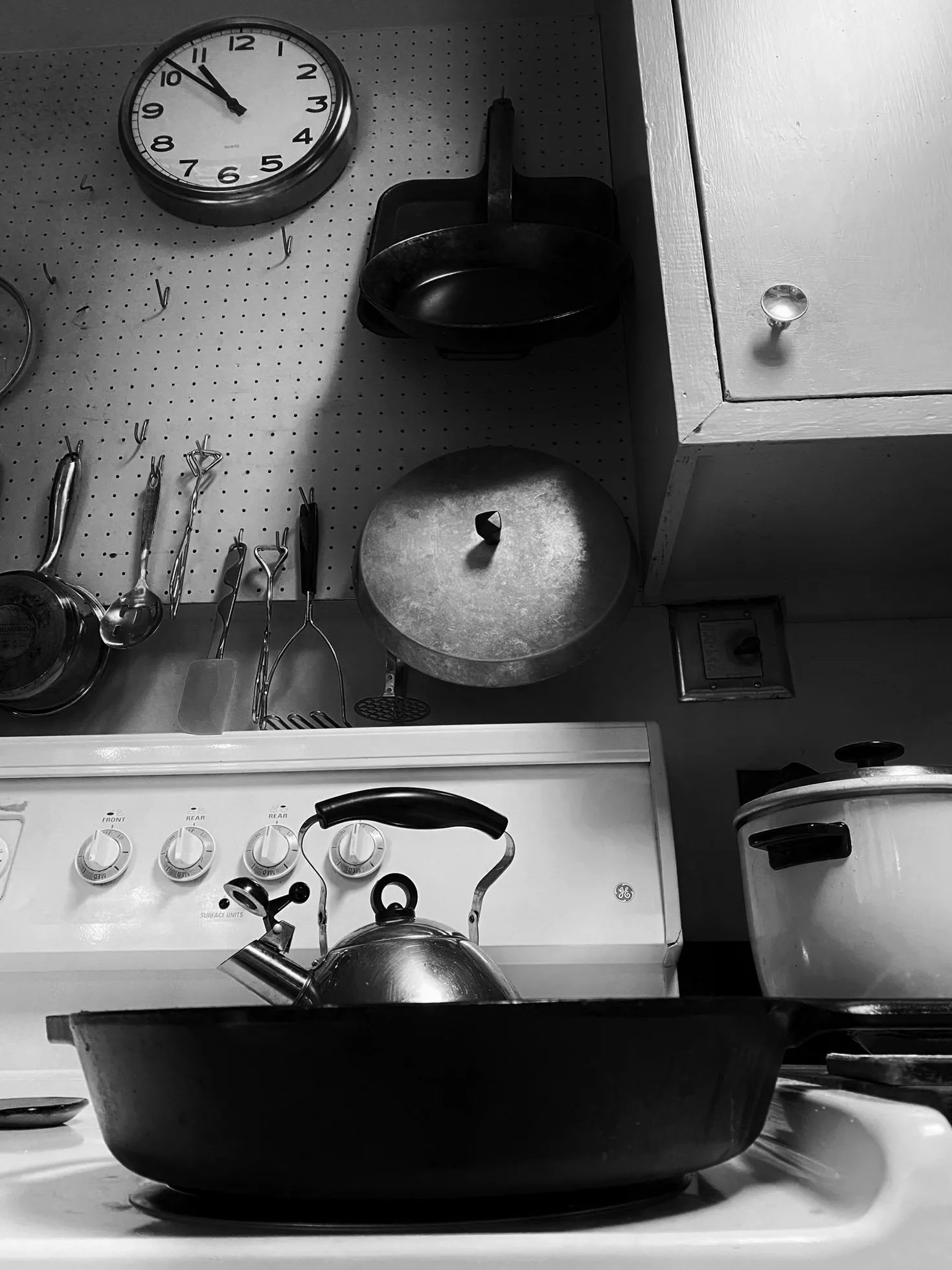 A kitchen with a pan in front of a teakettle on a white stove that is under a useful, large pegboard covering the wall from which utensils and pots and pans hang at the ready. There is a utilitarian clock on the wall indicating it is 10:52.