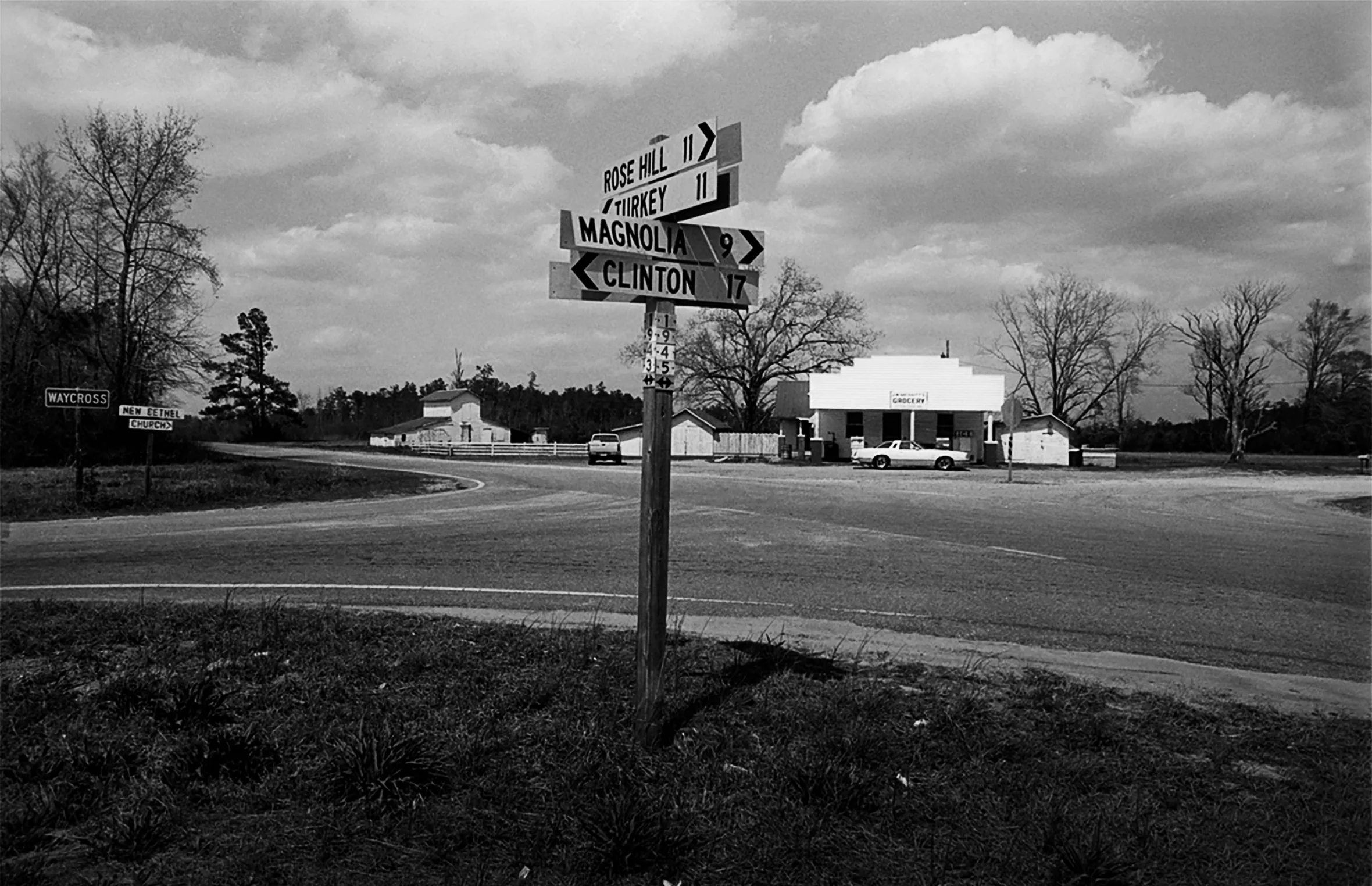 Crossroads signage in Duplin County, North Carolina, in 1989: Rose Hill, 11 miles; Turkey, 11 miles; Magnolia, 9 miles; and Clinton, 17 miles. In the background is a roadside grocery store with an older model car.