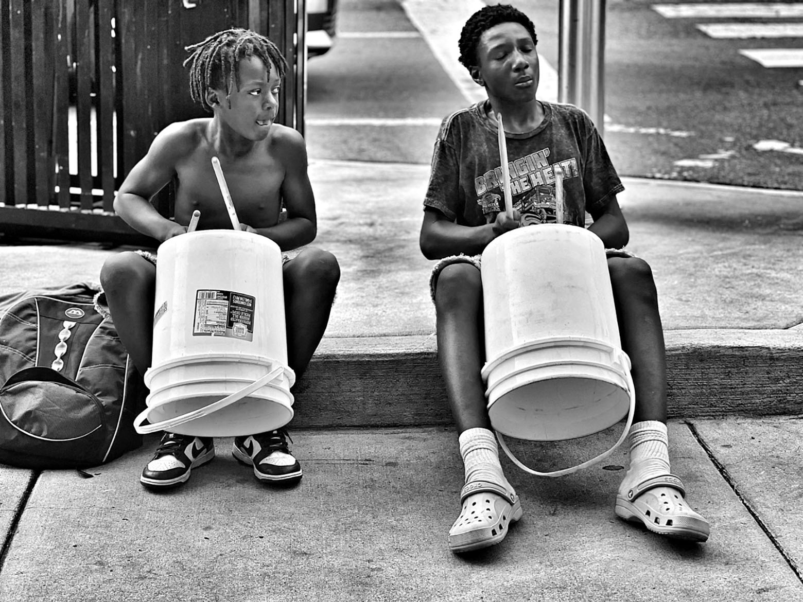 Two Black youths playing drums made from buckets on sidewalk. Interior photo for Readers Write on 