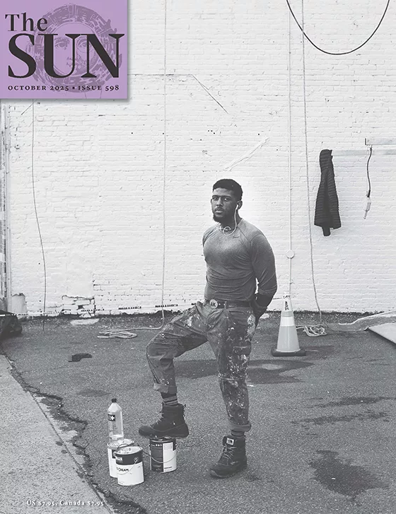 A Black artist about to paint a mural stands with his foot on a paint can in front of a white brick wall