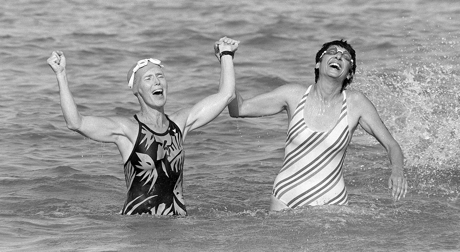 Two women swimmers standing in the ocean raise one arm up together in joyful triumph