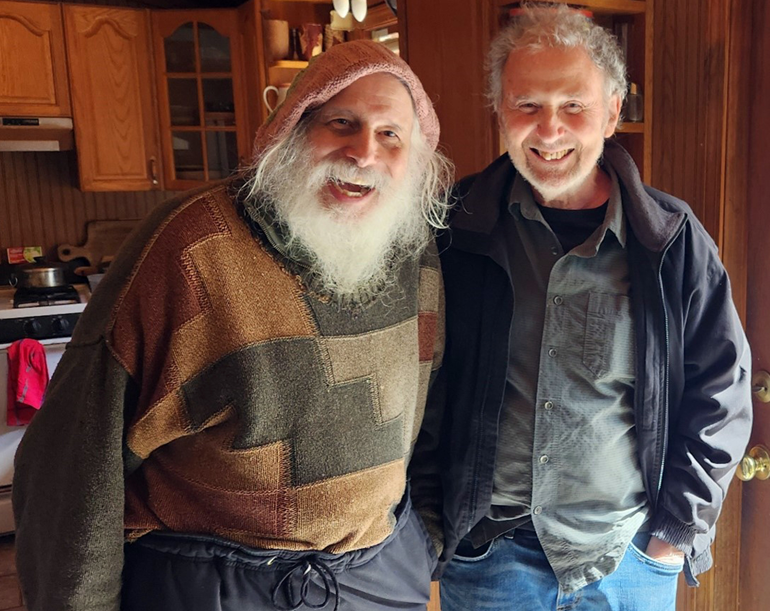 Sparrow and Sy Safransky are smiling and standing side-by-side in Sparrow's double-wide in Phoenicia, New York.