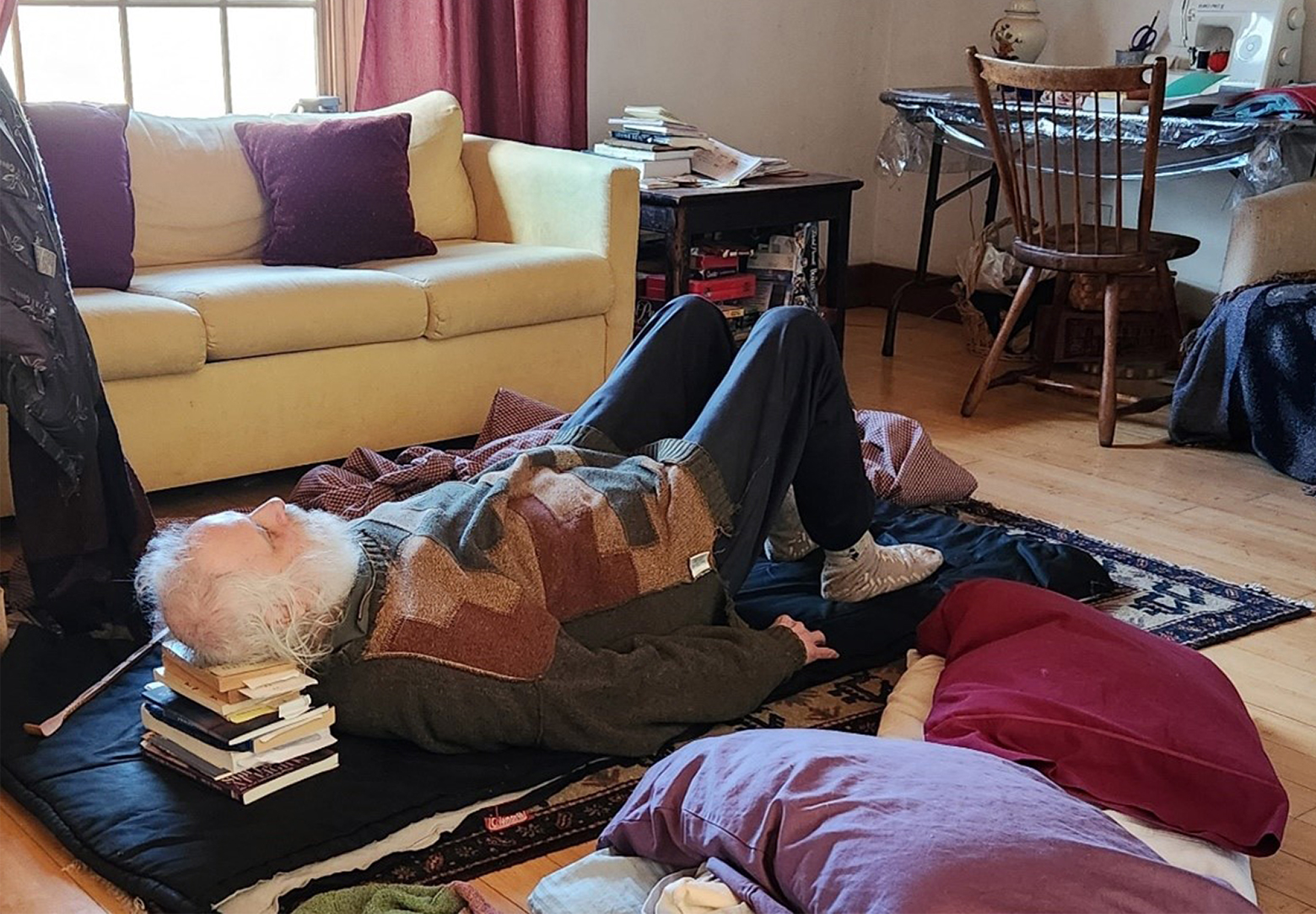 Sparrow is on his back, on a pad and a carpet on the floor with pillows strewn about. A stack of poetry books elevates his head. His arms rest on the pad with hands palms down, and his stockinged feet are flat on the floor and his knees are elevated.
