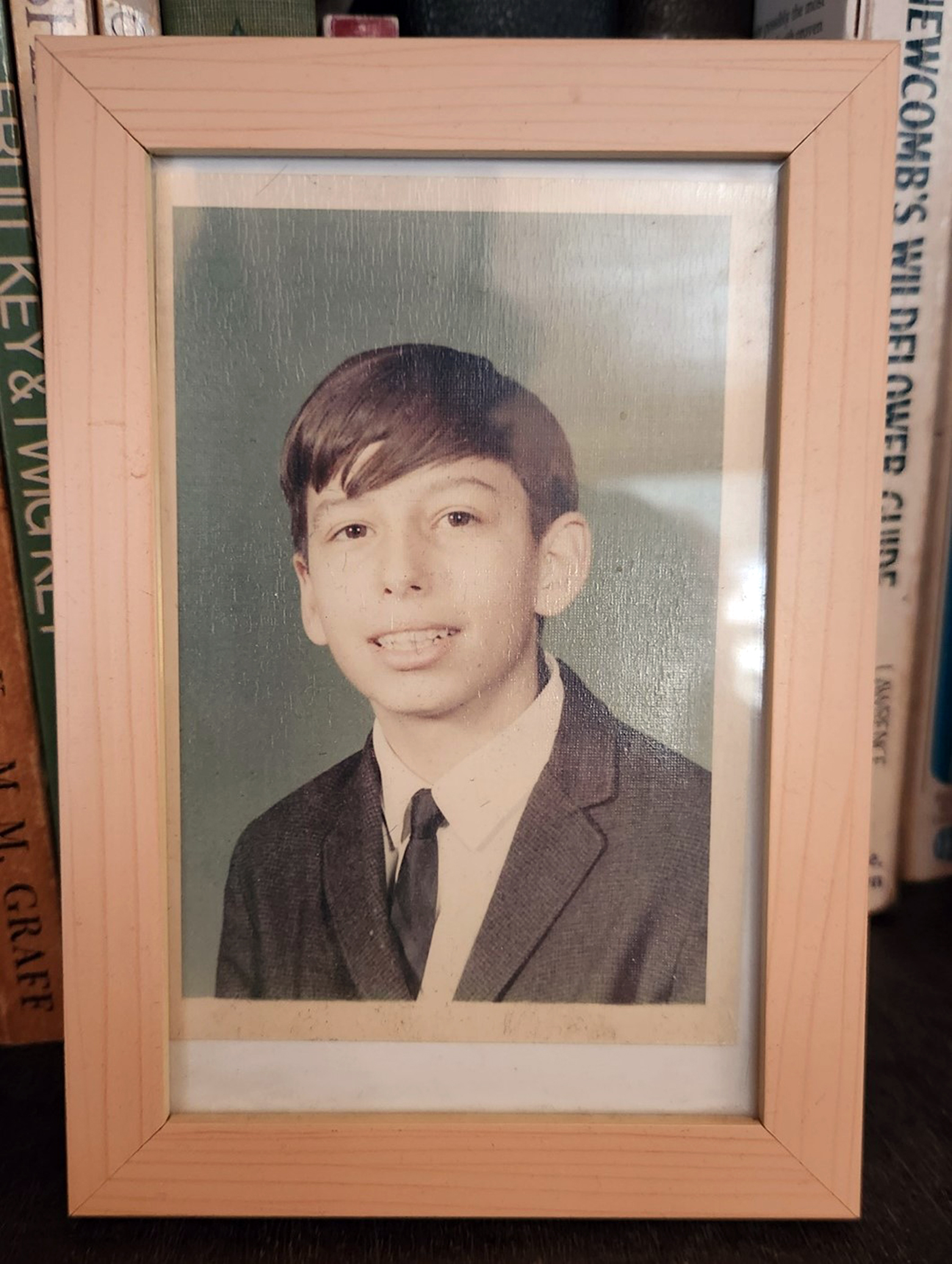 A wooden-framed school photo of Sparrow sits on a bookshelf in front of Newcomb's Wildflower Guide. Sparrow looks to be in grade school, and he is wearing a suit and tie.