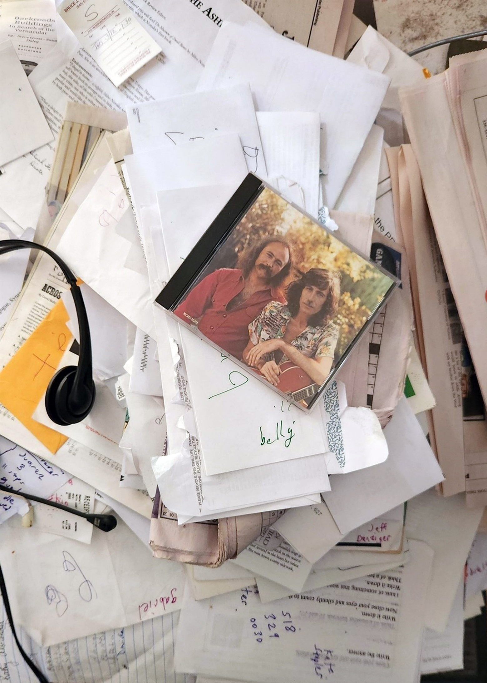 Sparrow's desk with mounds of unkempt papers and envelopes with a headset and a CD case featuring David Crosby and Graham Nash on top.