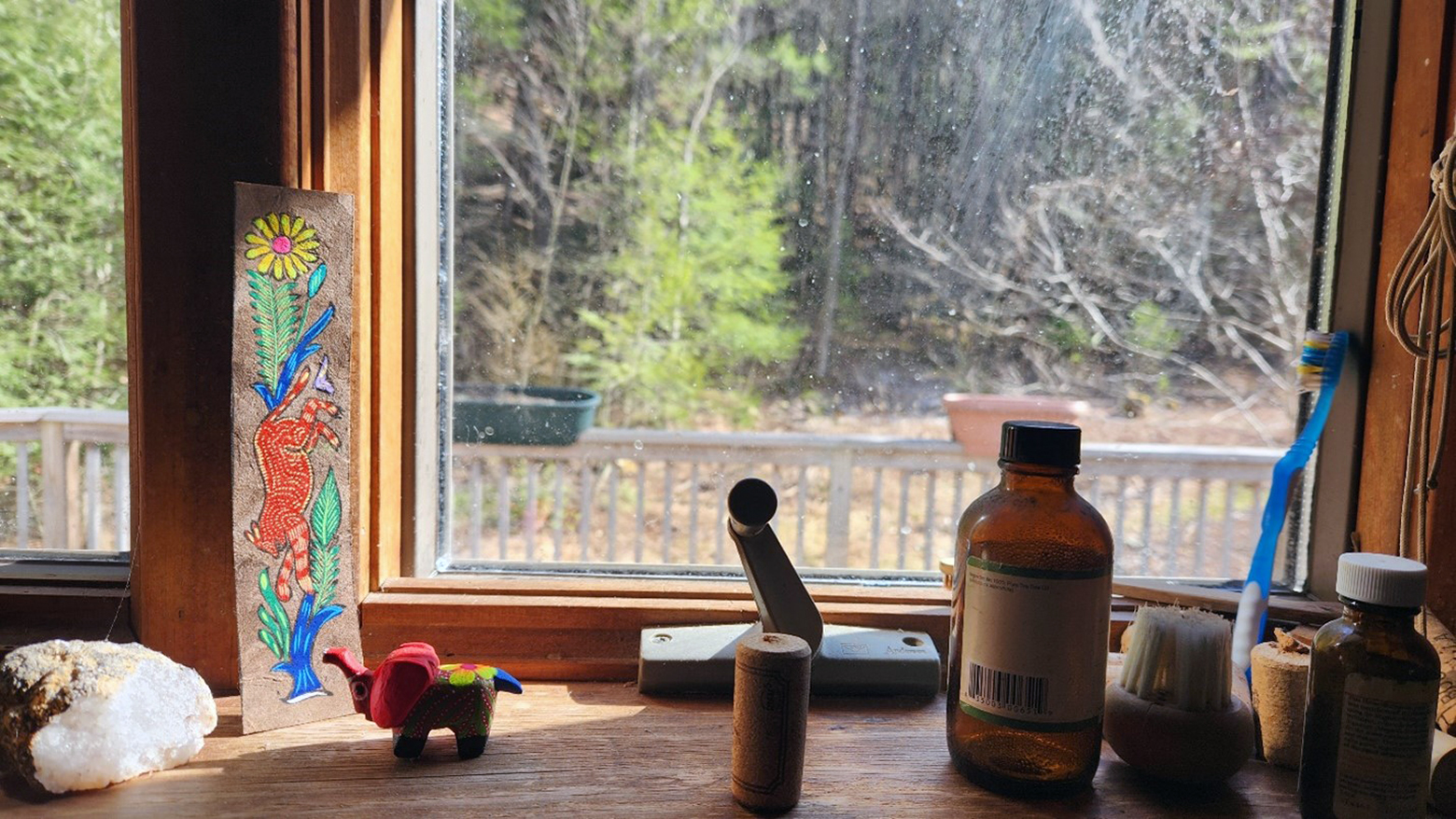 A window looks out on Sparrow's wooded backyard. On its ledge are a crystal; a narrow, vertical painting on wood board with a flower, butterfly, wild animal, and bird; a small, colorful elephant; a wooden spool; and  an empty glass medicinal bottle.