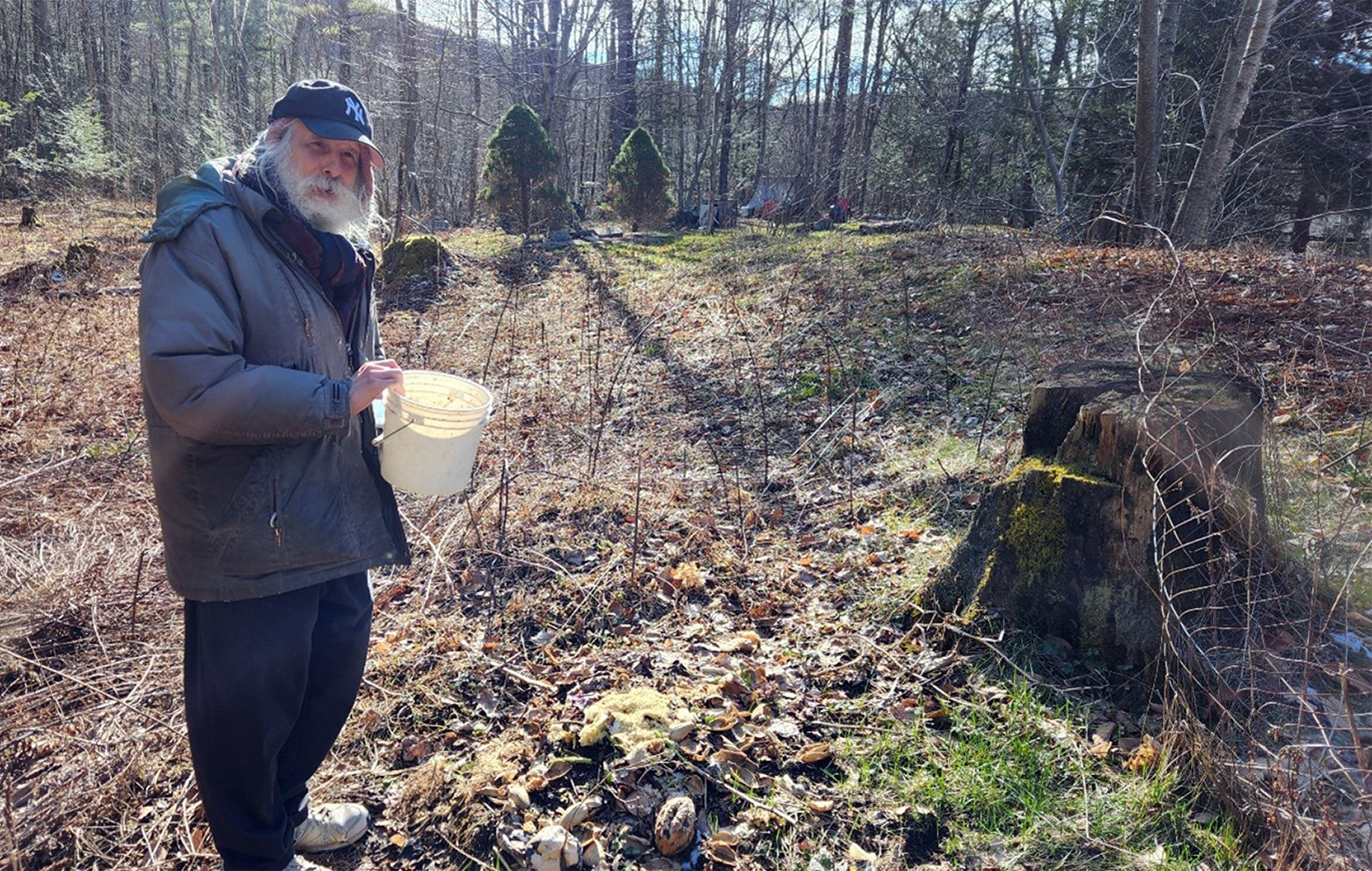 Sparrow stands in his wood-fringed backyard holding a white pail above a mound of debris on the ground in front of a large tree stump. He is wearing a ball cap with N-Y on it, a scarf and puffer coat, sweatpants, and tennis shoes.