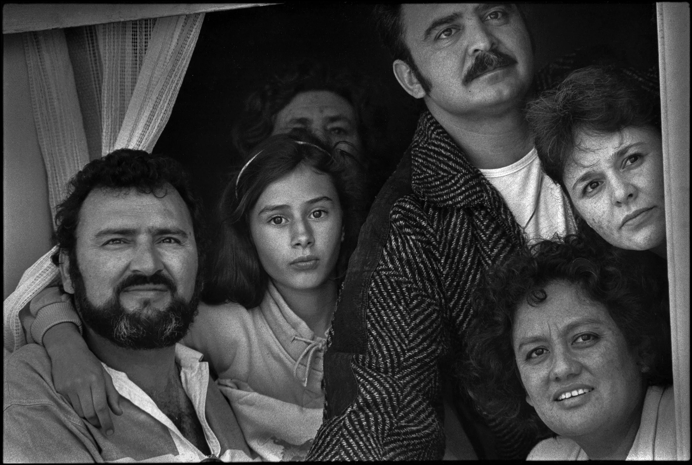 Six family members—two men, three women, and one girl—watch the annual San Francisco Carnaval Grand Parade in 1989 from their apartment window in the Mission District. Some members are smiling; all are looking out intently.
