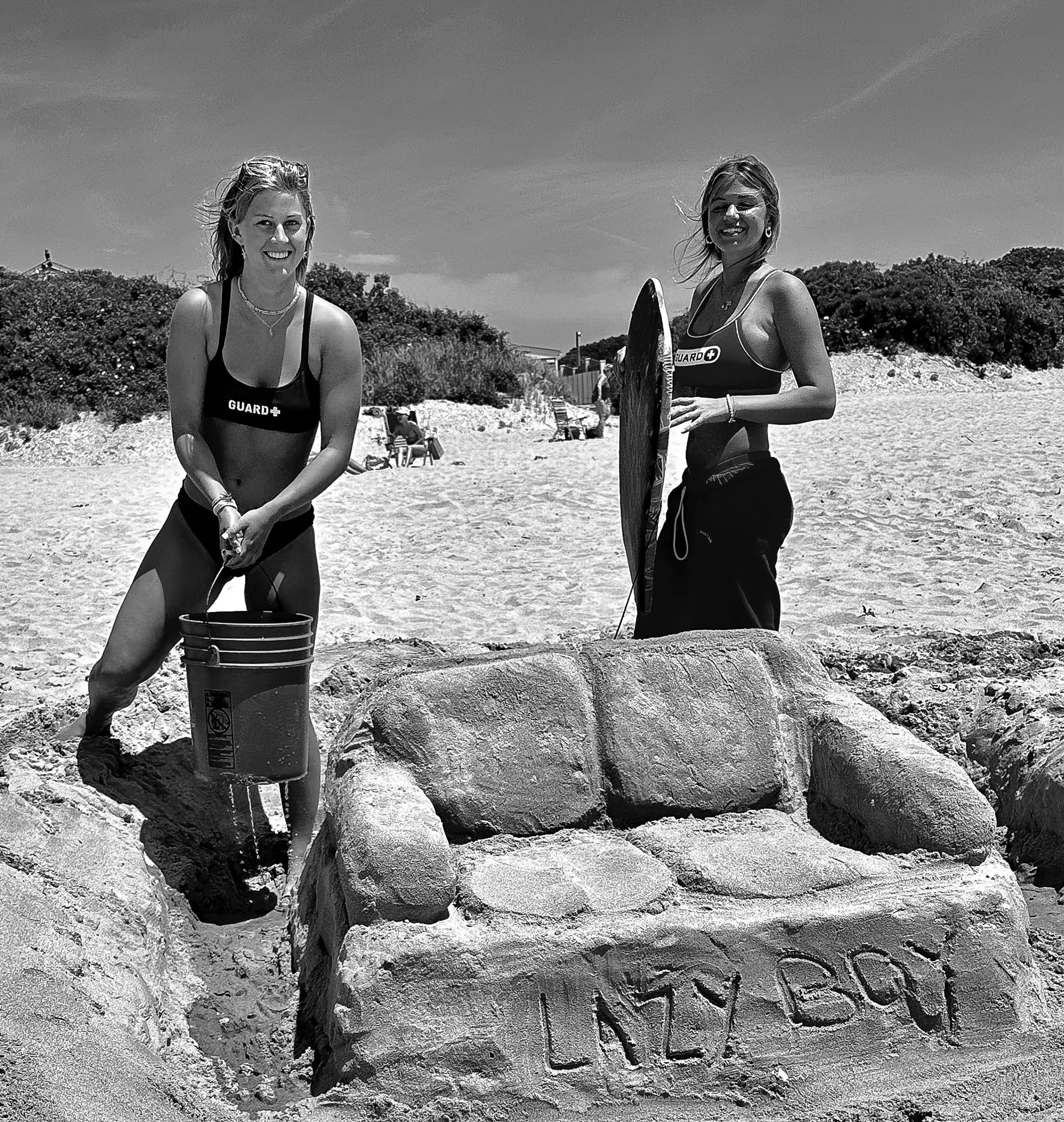 Two female lifeguards stand by a "Lazy Boy" couch made of sand