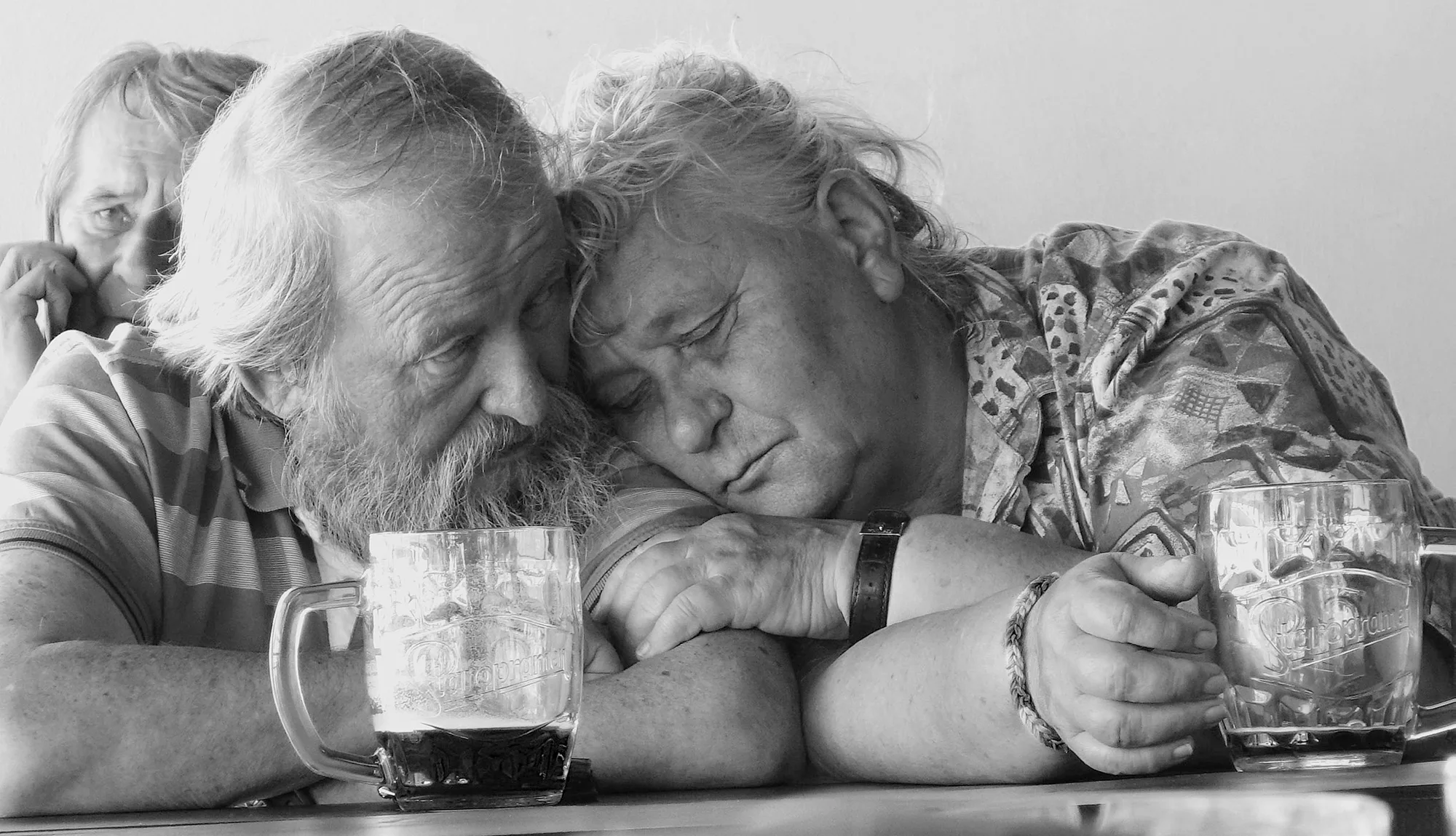 A woman with eyes closed and a beer in hand leans into a man seated next to her 