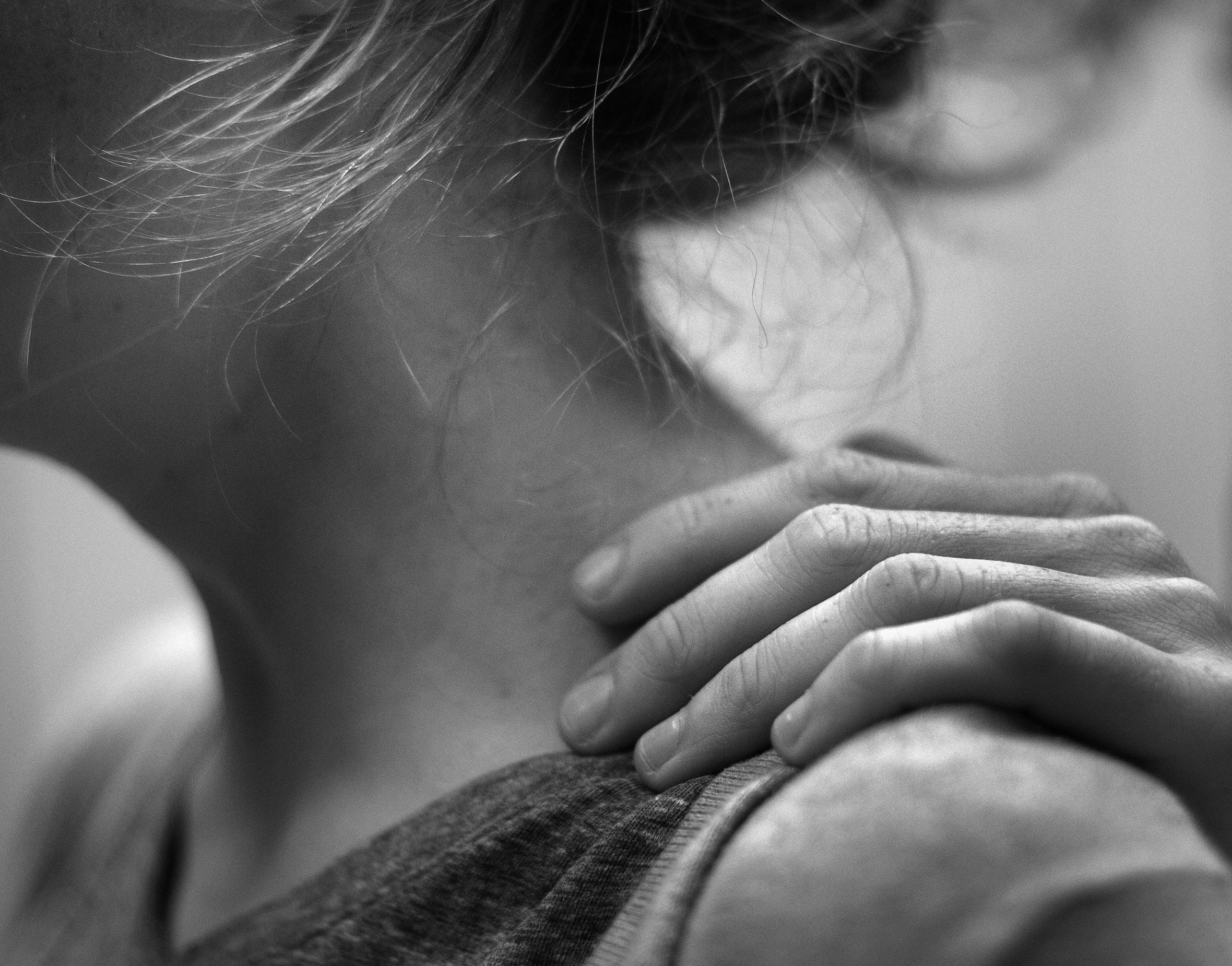 A close-up of the neck and shoulder area of a woman in a tank top with upswept hair facing left. A hand from behind rests on her left shoulder.