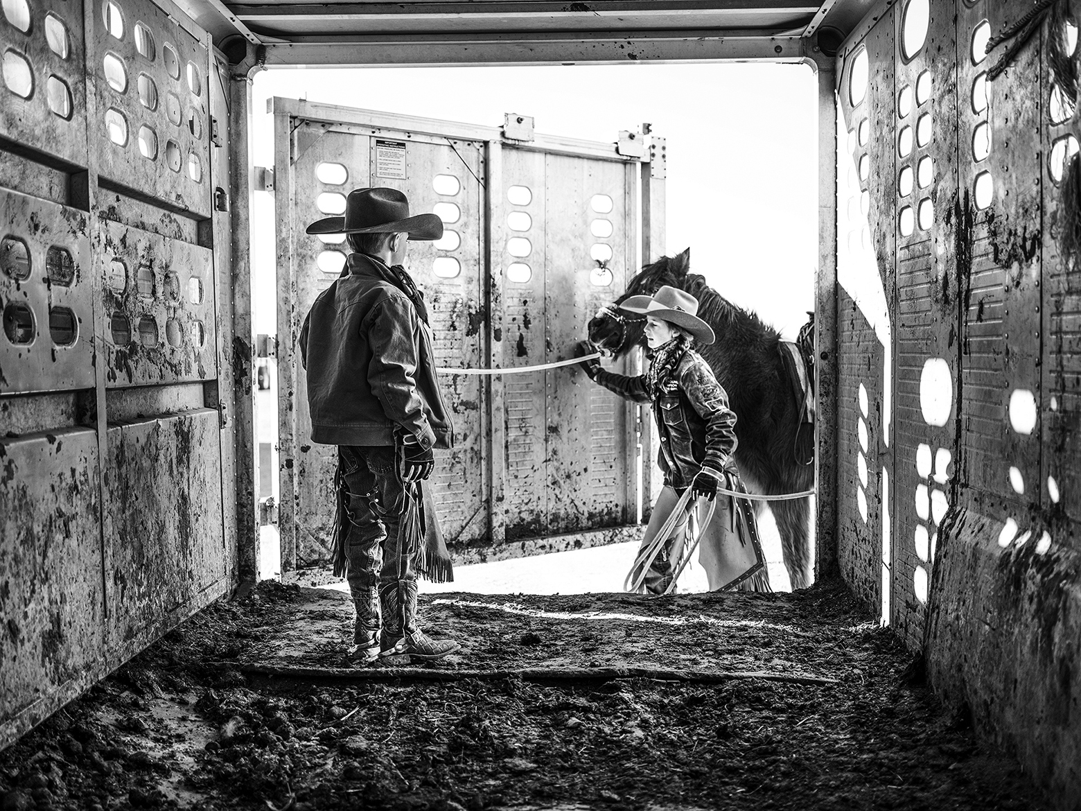A boy is waiting in a horse trailer as a girl guides a horse into it. Both are wearing cowboy hats and other typical Western wear.