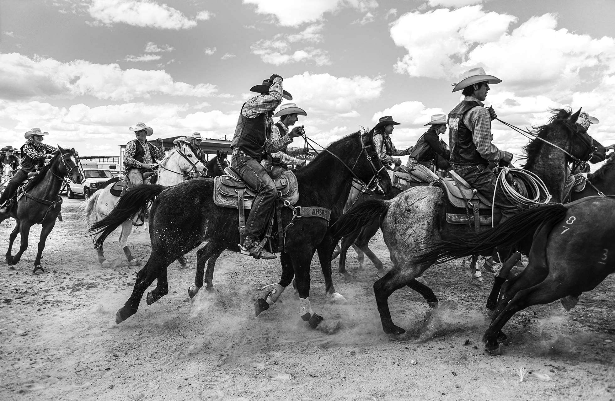 A group of men and women on horses with Western hats and jeans gallop toward the right of the image.