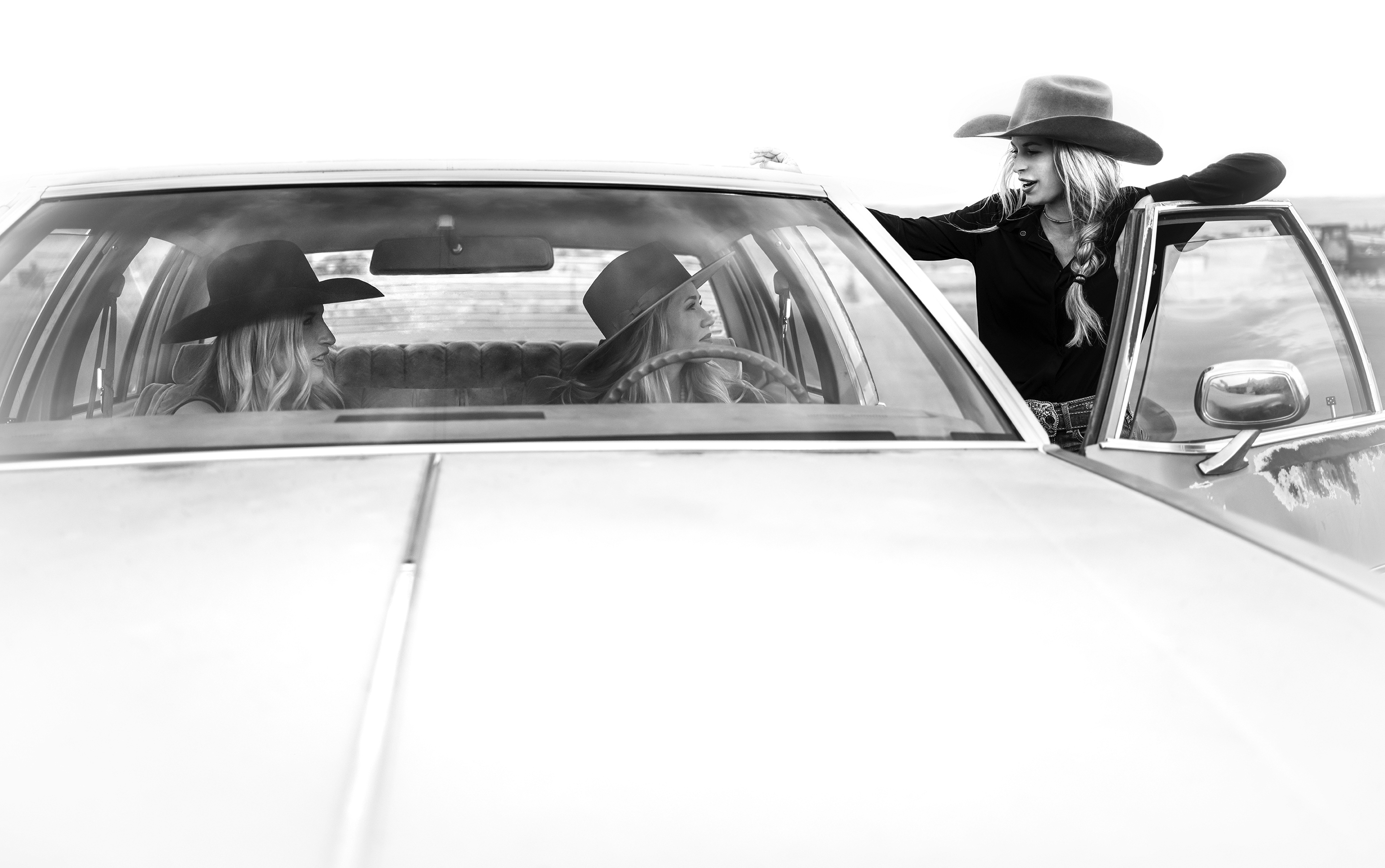 Two women in cowboy hats sit in a car and look to the right where another woman in a cowboy hat stands and leans against the open driver side door.
