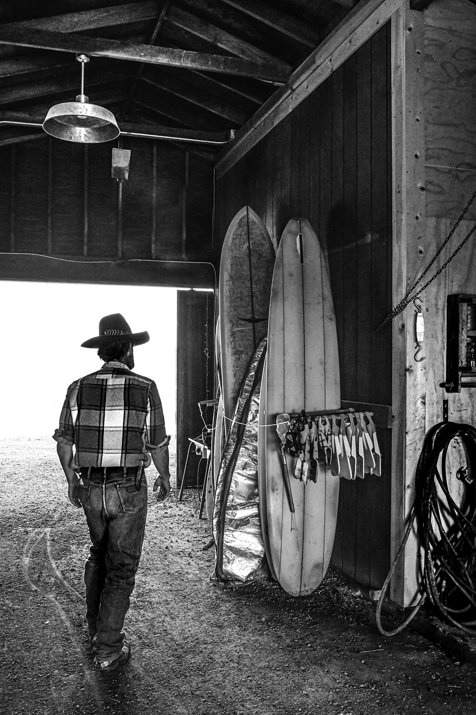 A man in a cowboy hat, plaid shirt, and jeans stands in a barn.