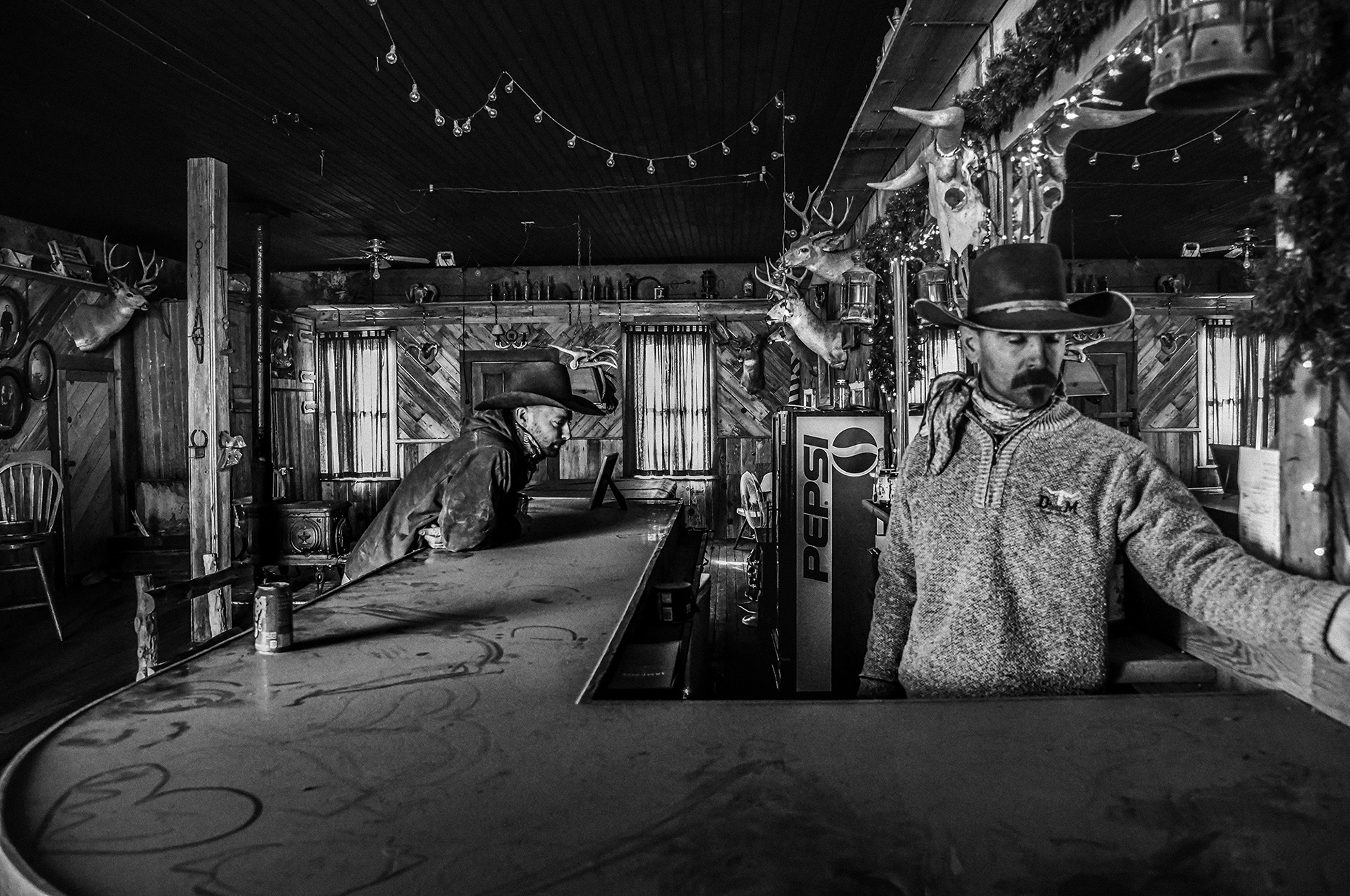 A bartender stands at the front. At the side counter, a man leans forward. The bar is covered with deer heads, wood paneling, and Christmas lights.