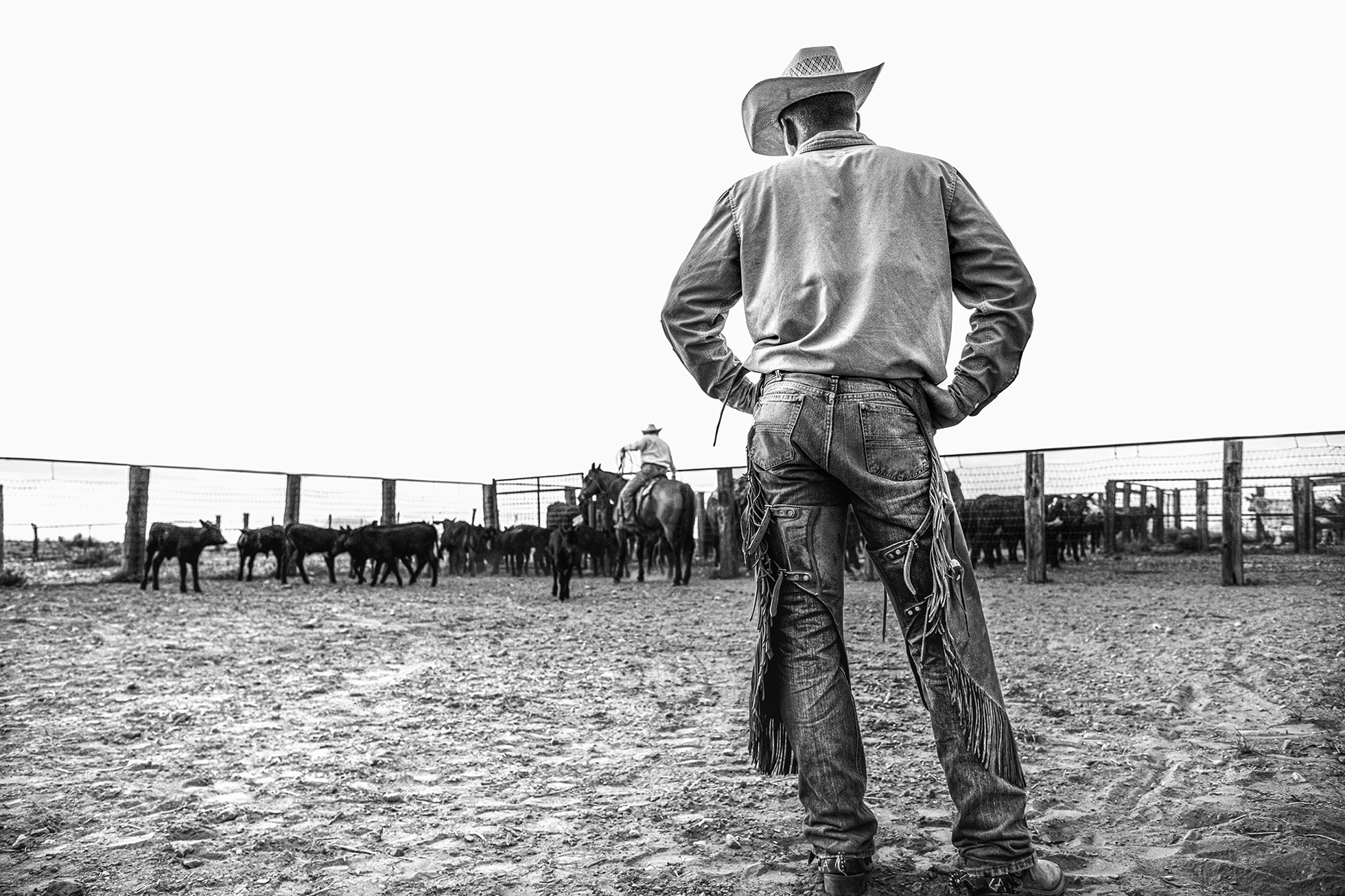A man with his back to the camera in jean shirt, denim pants, and a cowboy hat stands with his hands on his hips as he watches someone on a horse in the distance.