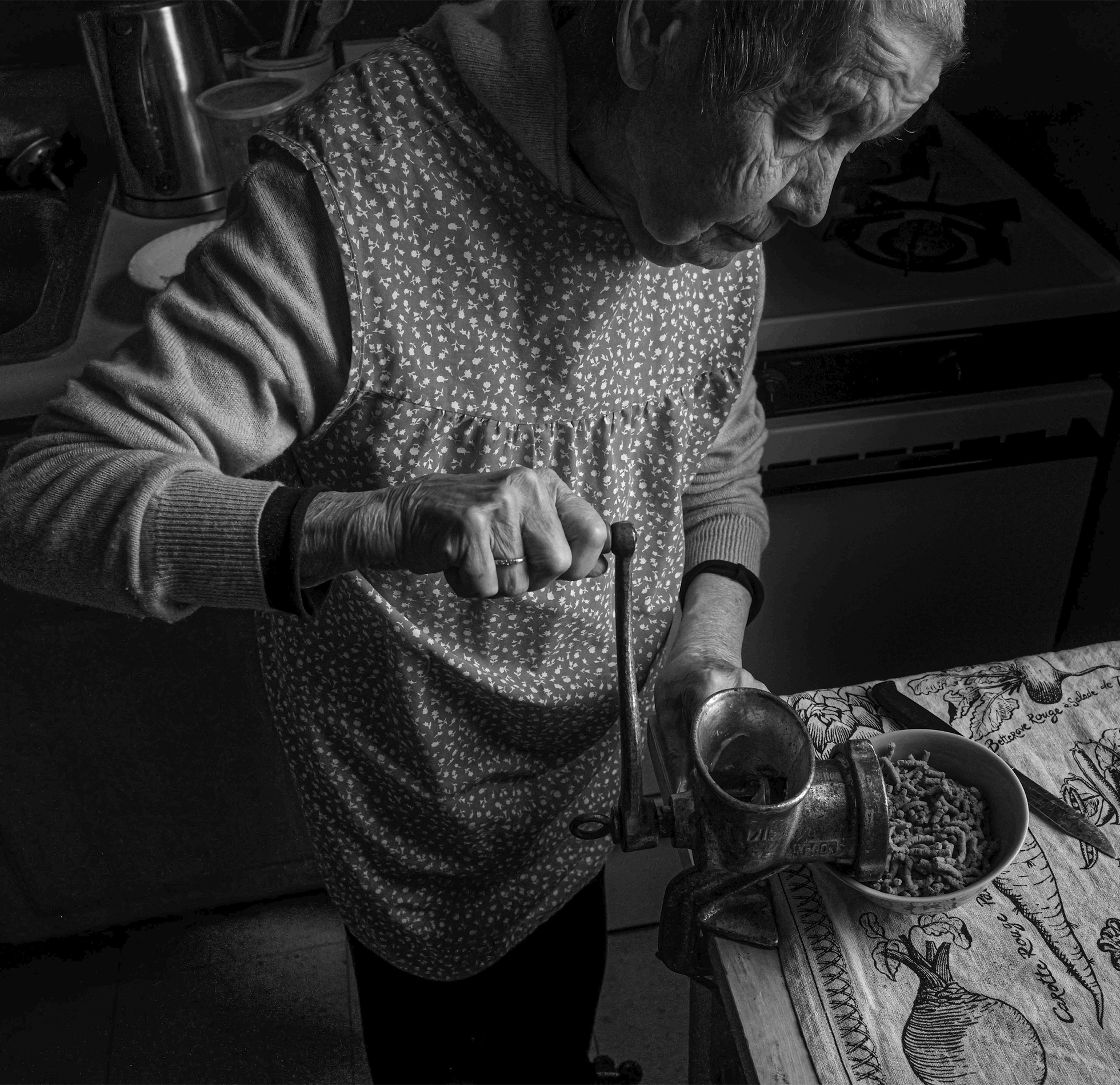 An old woman, with an apron over her sweater, stands at a kitchen table hand-grinding meat. The meat grinder is attached to the edge of the table.