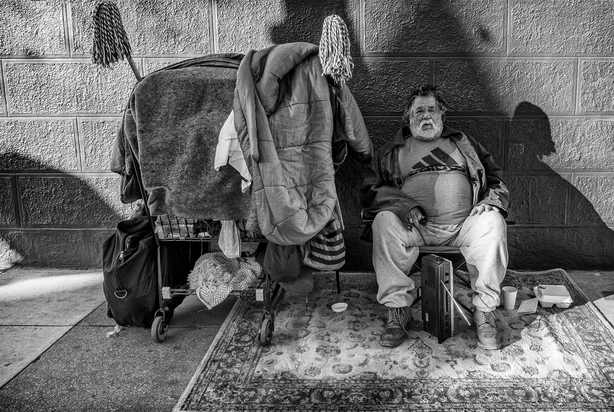Charlie sits on a chair with a large carpet underneath on a sidewalk in front of a wall with his belongings in a shopping cart on his right in San Francisco in December 2019.