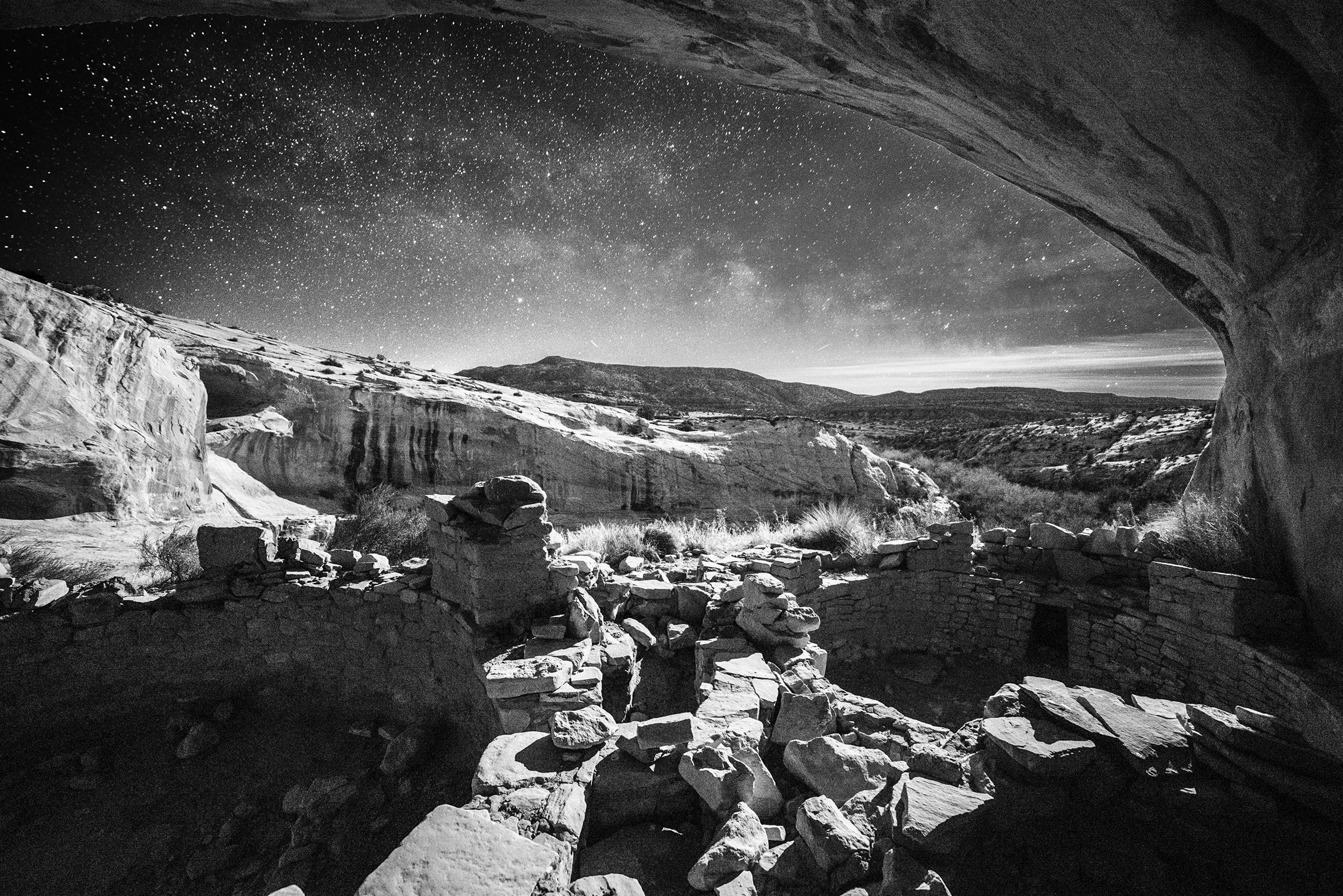 The photo titled “Ancient Nights: Sky Kiva” shows Anasazi ruins on a starry night. The ruins are shot from inside looking out of an overhang in a cliff with a rounded, stone-layered, short wall surrounding an area dug into the ground.